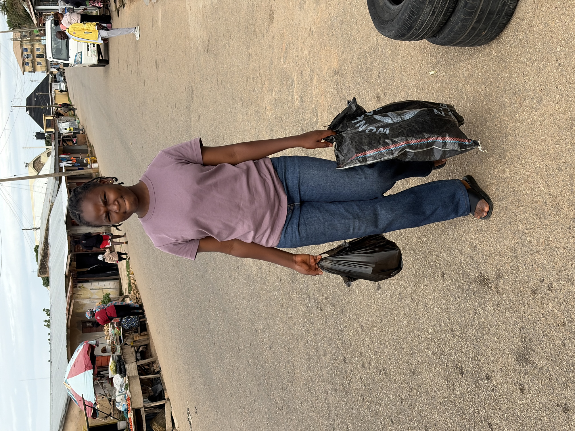 A young Black female with braided hair and a purple t-shirt and blue jeans walks on a paved road, carrying two large black plastic bags, one in each hand. Her expression is one of mild displeasure. In the background, along the side of the road, are several market stalls with corrugated metal roofs, selling produce and other goods. A few people are visible browsing or working at the stalls. Further down the road, a white van and a man in a yellow jacket are seen. The scene appears to be a street market in Jos, Nigeria, with the typical urban environment including buildings and a road. There are also two stacked tires on the right side of the image.