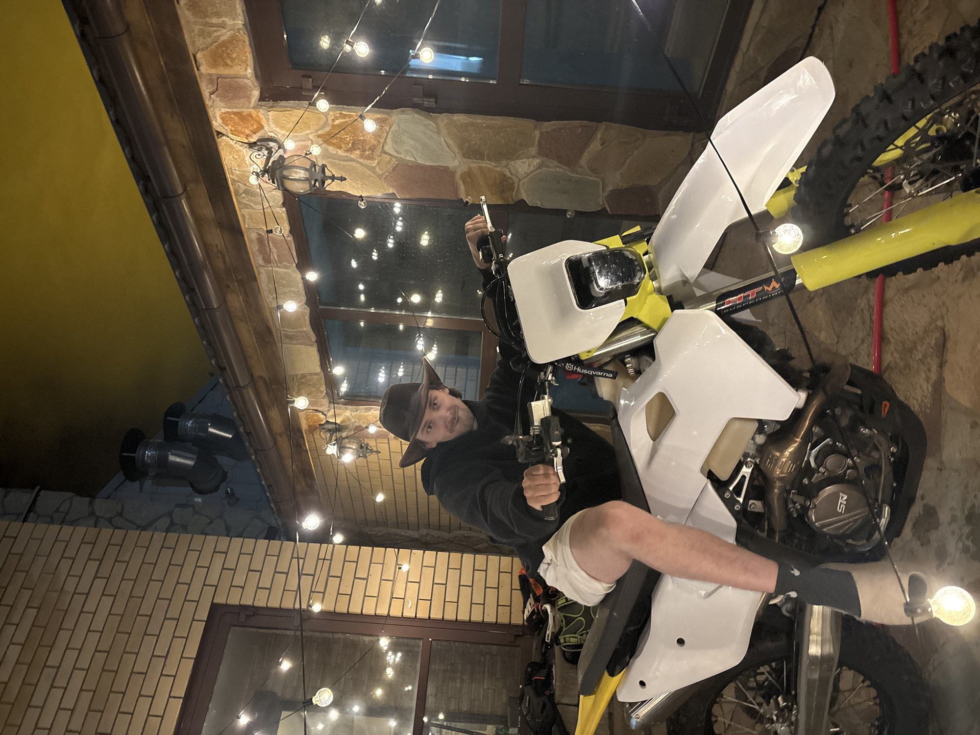 An outdoor evening scene in Ореховка, Russia, features a young man posing on a white and yellow dirt bike. He wears a dark, wide-brimmed hat, a black long-sleeved top, white shorts, and black socks. Seated on the motorcycle, he holds a black pistol in his right hand, pointing it slightly forward, and looks towards the camera with a slight smirk.
The motorcycle is a Husqvarna model, with "Husqvarna" visible on a strap near the handlebars. Other text includes "HTM Suspensions" on the front fork and "SR" on the engine casing.
The background reveals a textured building facade, composed of both stone and brick walls, along with large windows that reflect the ambient light. A dark wooden beam stretches across the upper part of the scene. An outdoor light fixture is mounted on the stone wall, and metal vents are visible on a roof section. Numerous glowing spherical string lights are strung horizontally across the frame, providing warm illumination for the nighttime setting.