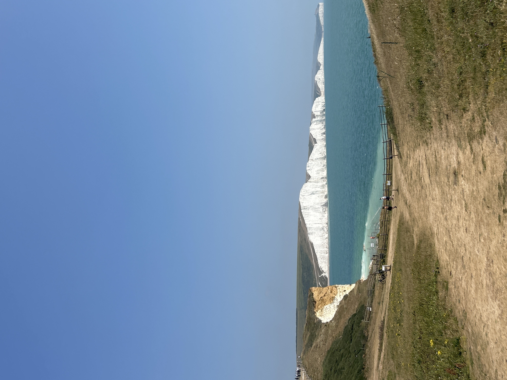 A wide shot captures the Seven Sisters cliffs near Seaford, United Kingdom, on a clear, sunny day. The bright blue sky dominates the upper half of the frame. The sea, a vibrant turquoise, stretches out in front of the majestic white chalk cliffs. The cliffs are irregular in shape, with some areas appearing to have eroded. In the foreground, a dirt path winds through dry, golden grass. A wooden fence runs along the edge of the path, separating it from a sloping grassy area. Several people are visible on the path and on the cliff edge, some walking and others standing. Two bicycles are parked near the fence. In the distance, the white cliffs extend along the coastline, gradually fading into a hazy horizon. The overall impression is one of a scenic coastal landscape with a few visitors enjoying the view.