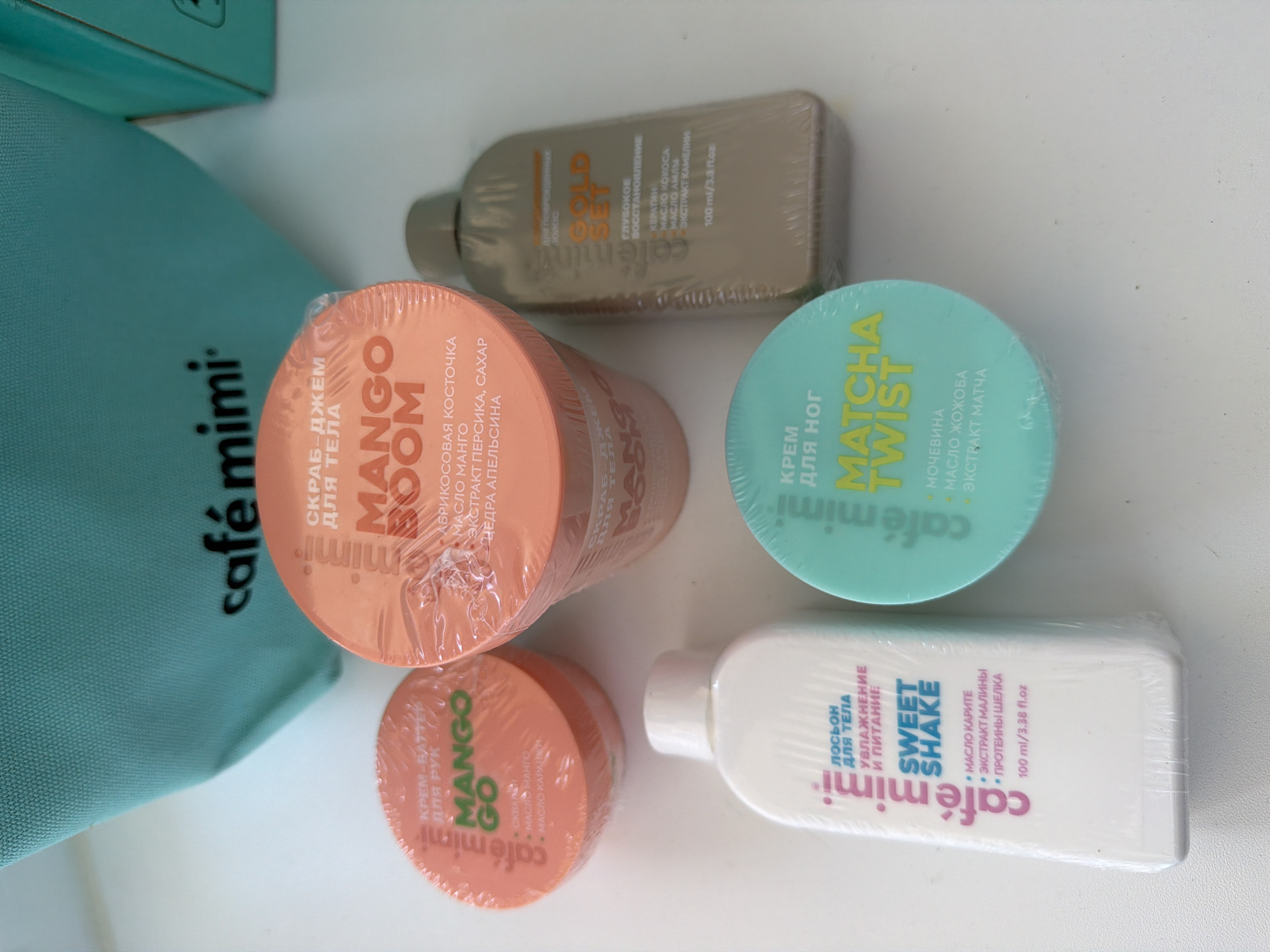 Here is a detailed description of the image:
The image shows a collection of skincare products arranged on a white surface. The products are from the brand "café mimi."
From left to right and top to bottom, here's a description of each item:
1. **Café mimi bag**: The left side of the image includes part of a turquoise bag with the "café mimi" logo in black.
2. **Gold Set**: At the top, there's a tan-colored bottle labeled "SOLD SET", with a description of "Deep Recovery" (translated from Russian) and key ingredients like keratin, coconut oil, amla oil, and camellia extract. Its volume is 100 ml/3.8 fl.oz.
3. **Mango Boom**: The next item down is a round, orange container labeled "MANGO BOOM" body scrub jam with the ingredients apricot kernel, mango oil, peach extract, sugar, and orange zest.
4. **Matcha Twist**: Next to Mango Boom is a round, light blue container labeled "MATCHA TWIST" foot cream, containing urea, jojoba oil, and matcha extract.
5. **Mango Go**: Below Mango Boom is a smaller round, orange container labeled "MANGO GO" hand cream butter, which contains mango oil and karashi oil.
6. **Sweet Shake**: At the bottom right, there's a white rectangular bottle labeled "SWEET SHAKE" body lotion, promising moisturizing and nourishing properties. Its key ingredients are shea butter, raspberry extract, and silk proteins. Its volume is 100 ml/3.38 fl.oz.
Most of the products are covered in a clear plastic wrap, which suggests they are new or unused. The arrangement of items gives the image a feel of being a product display or a collection of items laid out for review or use.