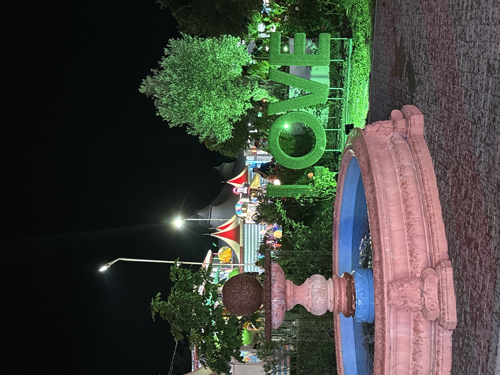 This is a nighttime photograph of an outdoor attraction in Kabardinka, Russia. In the foreground, a pink, multi-tiered fountain with a spherical top is partially submerged in a blue pool of water. To the right of the fountain, large, three-dimensional green letters spell out "LOVE." Behind the fountain and the "LOVE" sign, there are several structures with peaked roofs, illuminated by colorful lights, suggesting a carnival or amusement park setting. A large tree, illuminated with green light, stands behind the "LOVE" sign. The ground is paved with dark-colored stones. The sky is dark, indicating nighttime.