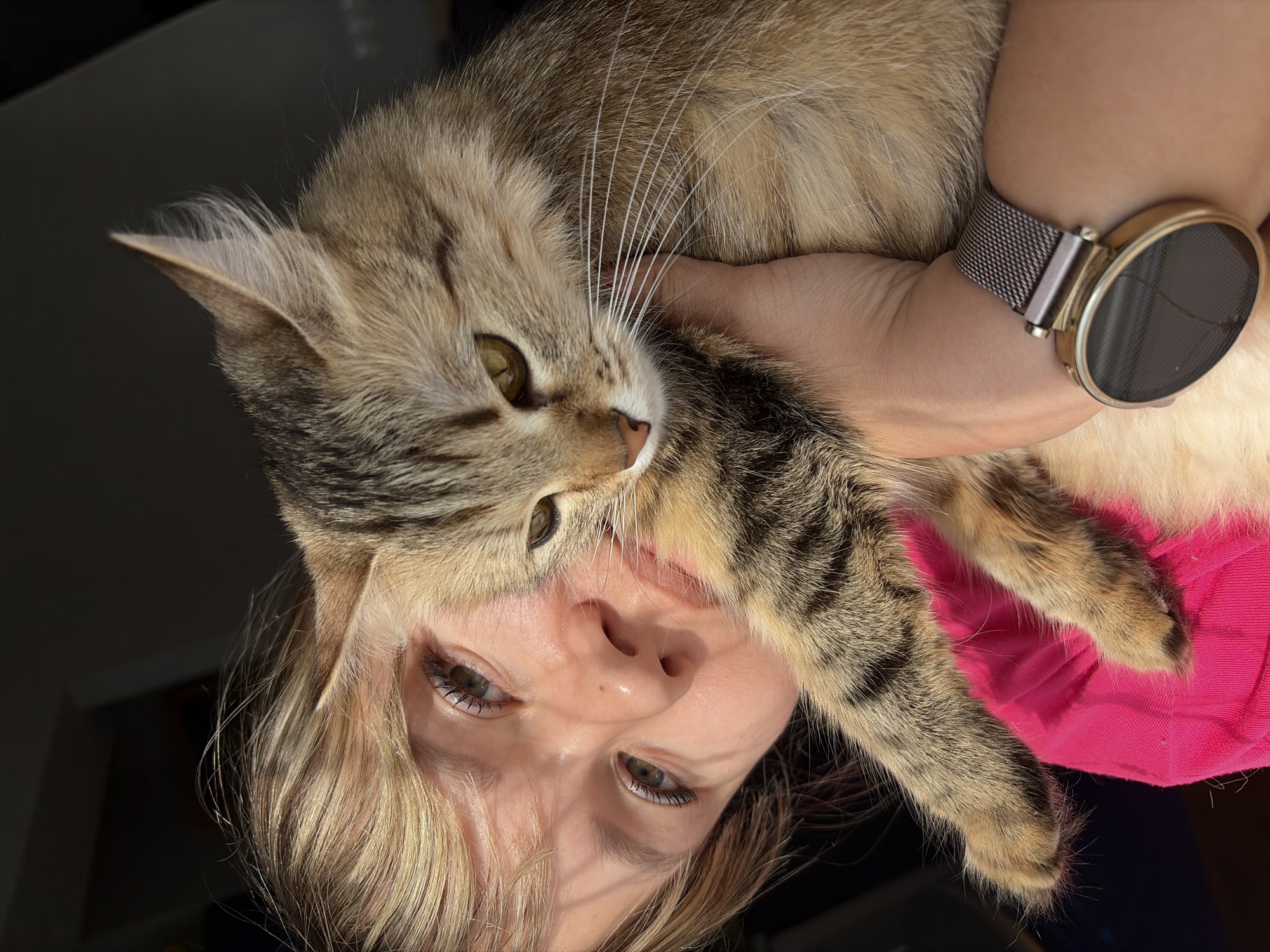 A woman is holding a cat, likely a pet, close to her face. The woman's face and the cat are the primary focus, filling much of the frame. The woman has light colored hair and is looking directly at the camera. She has a digital watch on her left wrist. The cat is light brown and tan with tabby markings. The background is dark and out of focus, with a hint of what might be a wall or furniture. A pink fabric is visible in the bottom right of the frame. The image orientation is rotated approximately 180 degrees. There is no information available in the image to confirm a location in Chaikovsky, Russia.
