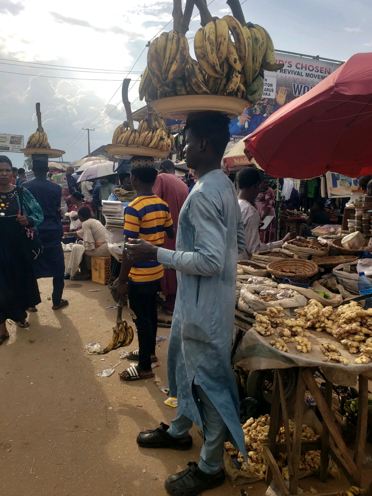 A medium shot captures a bustling outdoor market scene in Sabon Tasha, Nigeria. In the foreground, a young Black man wearing a light blue traditional garment stands facing right, his left hand resting on his side, while his right arm is bent and extended forward. To his left, another Black man, wearing a yellow and blue striped shirt and dark pants, faces left. He wears a patterned cap and has his hands clasped in front of him. Behind him, a Black woman in a dark dress with colorful accents stands facing the camera, her left arm slightly bent.
The market is filled with people in the background, some carrying items. Several banana bunches are prominently displayed, with two large bunches balanced on elevated trays, likely for sale. A red umbrella is visible in the upper right of the frame, partially obscuring a banner with text. In the mid-ground and right side of the image, tables are laden with goods, including large quantities of ginger root and what appear to be sacks of spices or grains. The ground is a dusty, light brown color. The sky above is partly cloudy with bright sunlight.