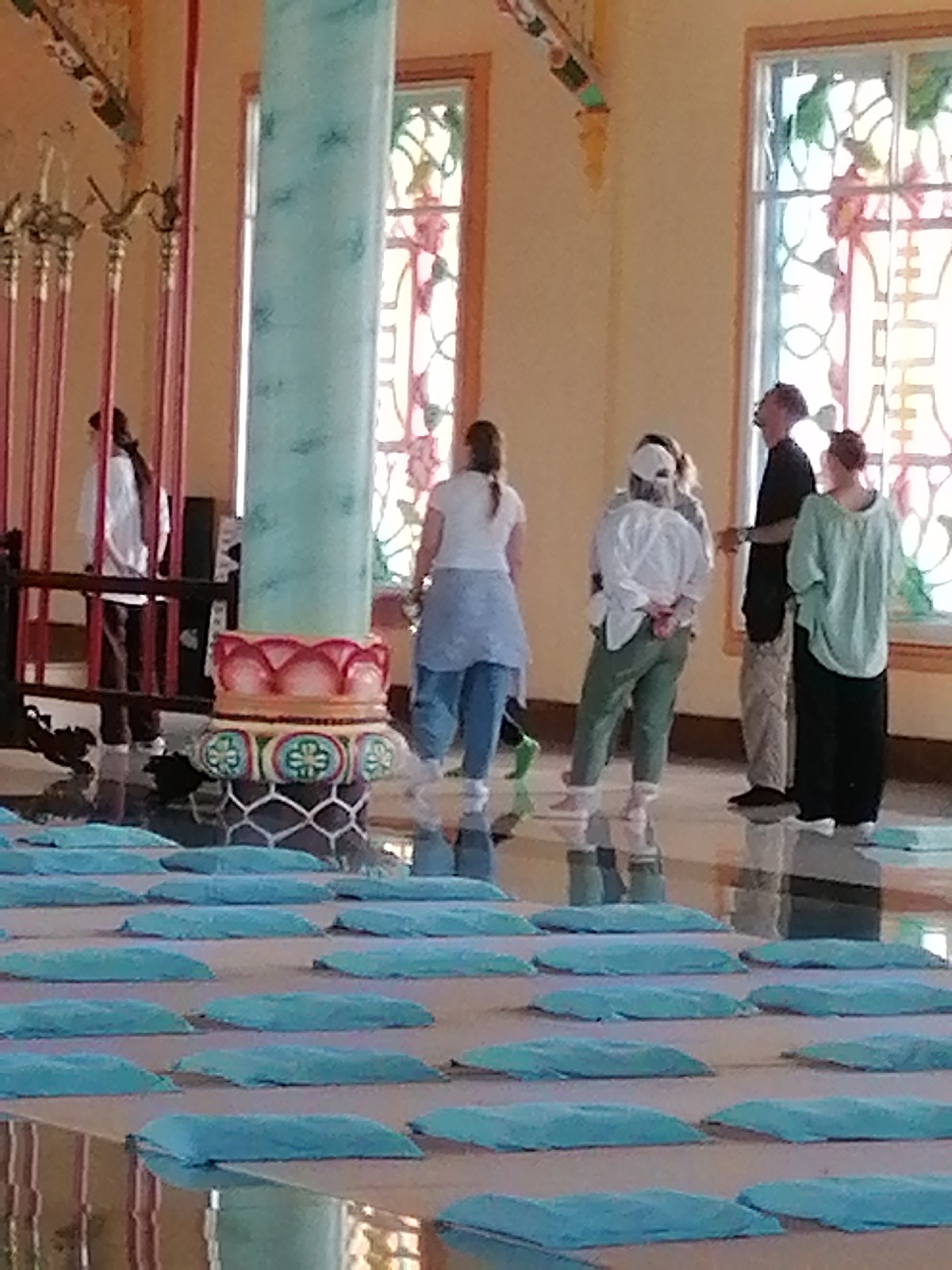 The image depicts the interior of a Buddhist temple in Lộc Quý, Vietnam. In the foreground, rows of light blue meditation cushions are laid out on the floor. In the middle ground, a large, teal-colored pillar with lotus-shaped decorative elements stands prominently. Several individuals are present in the background. One person, wearing a white shirt and dark pants, stands behind a red-barred enclosure. To the right of the pillar, three individuals are gathered: one person in a white top and green pants, another in a black shirt and light-colored pants, and a third in a light green top and dark pants. The background features tall windows with stained glass, casting colorful light into the space. The walls are a light, warm color, and decorative elements can be seen along the upper part of the walls. The floor appears to be reflective, showing a partial reflection of the individuals.