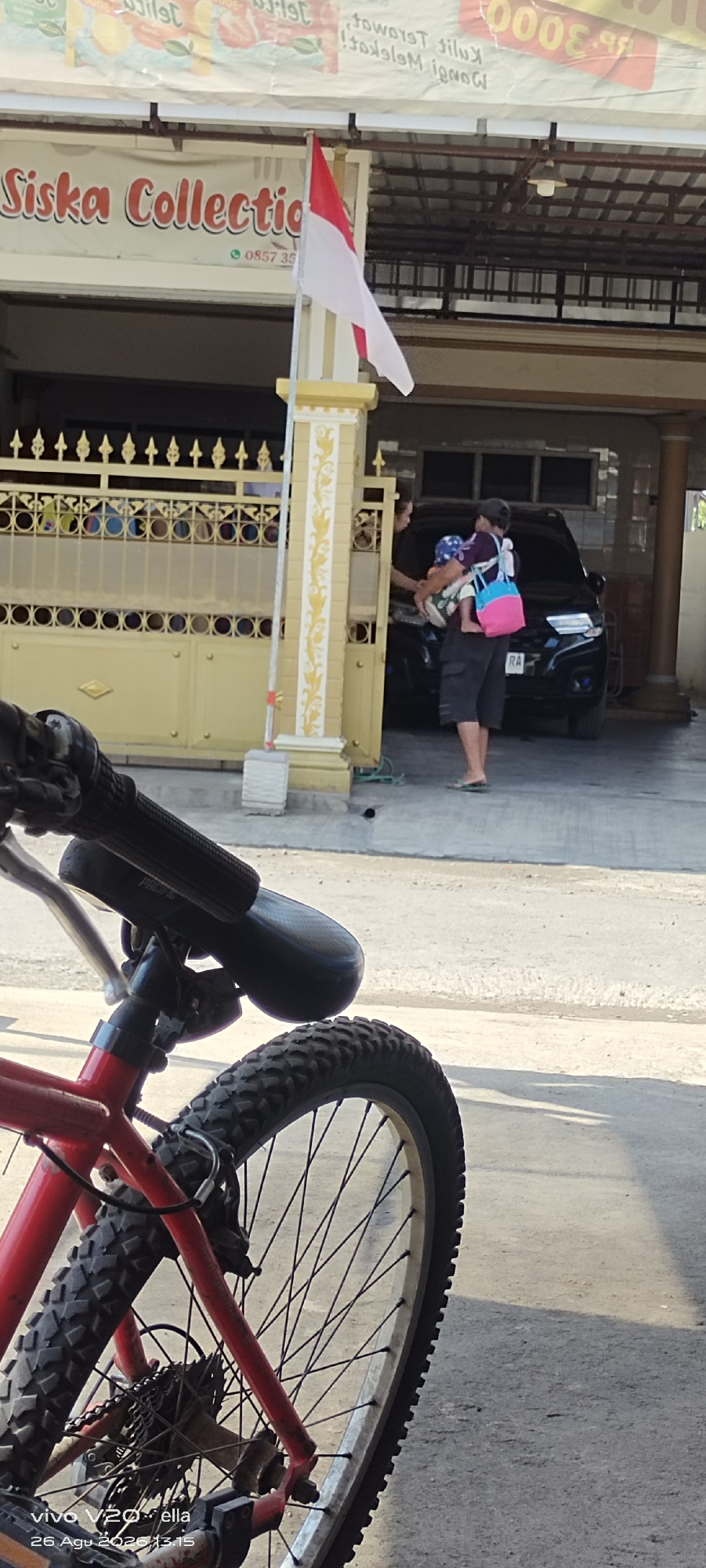 In the city of Simo, Indonesia, an Indonesian flag flies in front of a building with a sign that reads "Siska Collection." A person is seen holding a baby while standing next to a black car. In the foreground, a red bicycle is parked. The scene appears to be on a street with a concrete surface.