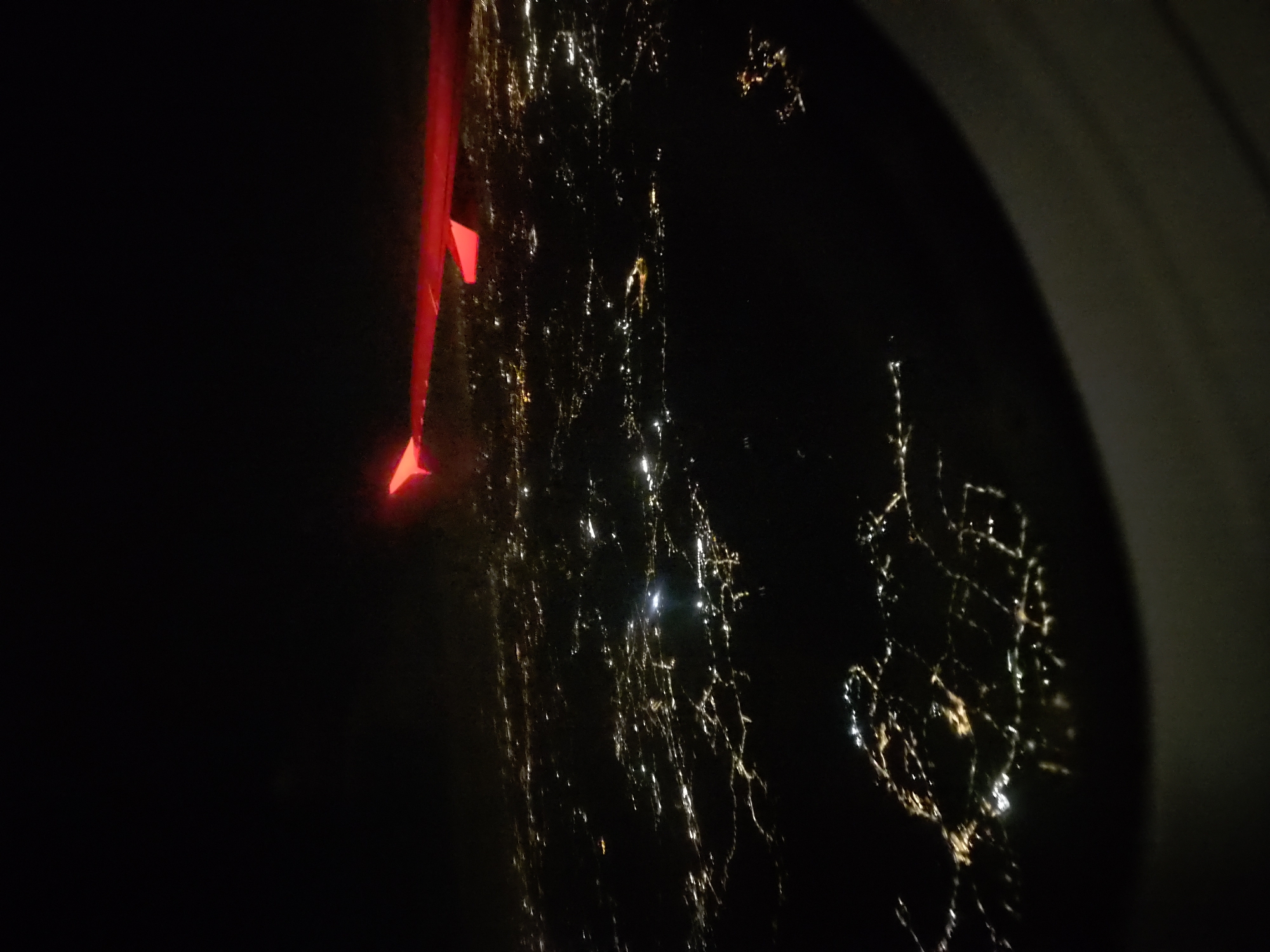 An aerial view captured from an airplane window at night reveals a sprawling cityscape illuminated by a multitude of lights. The red wingtip of the airplane is visible in the upper right portion of the frame, its bright color contrasting with the dark sky and the glowing urban landscape below. The city lights form intricate patterns, suggesting a network of streets and buildings, with some areas appearing denser and brighter than others, indicating commercial or residential centers. The overall impression is one of a vast, active metropolis under the cloak of darkness.