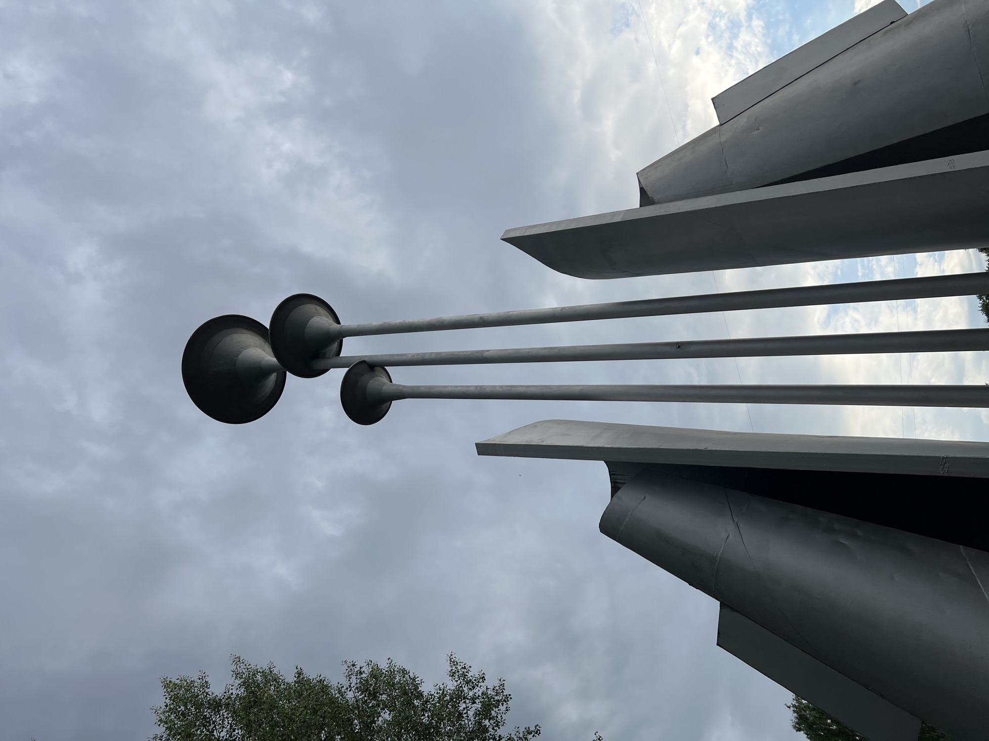 The image is an upward, low-angle shot of a modern art sculpture in Chaikovsky, Russia. The sculpture features three tall, slender poles topped with large, bowl-like lamps, positioned amidst large, dark grey, angular metal panels that form a dynamic, abstract structure. The sky above is overcast with dramatic, cloudy formations, with patches of blue sky visible. To the left of the frame, a portion of a leafy green tree is visible. The sculpture appears to be made of dark metal and is located outdoors.