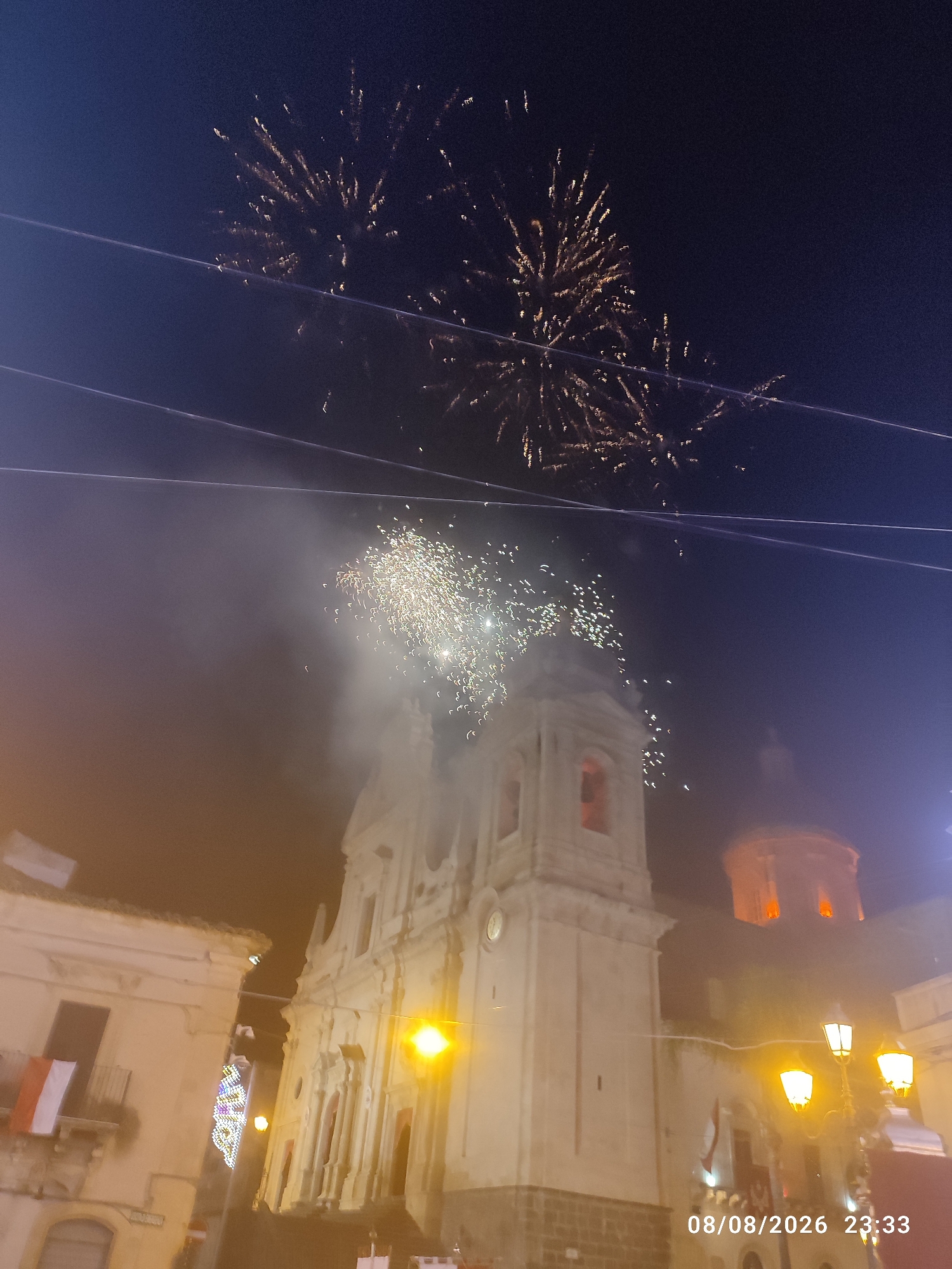 An outdoor nighttime scene captures a procession or festival in Militello in Val di Catania, Italy, with fireworks illuminating the dark sky above a town square. A large, ornate church, likely the Church of San Nicolò or another prominent religious building in the town, dominates the center of the frame, its bell tower and façade visible. Festive lights and decorations are scattered throughout the scene. Several flags, including one with red and white stripes, are displayed from buildings. The ground level appears to be paved, with streetlights casting a warm glow. The overall atmosphere suggests a celebratory event, possibly a religious feast or a local holiday, taking place in the historic center of Militello in Val di Catania. The image is dated 08/08/2026 at 23:33.