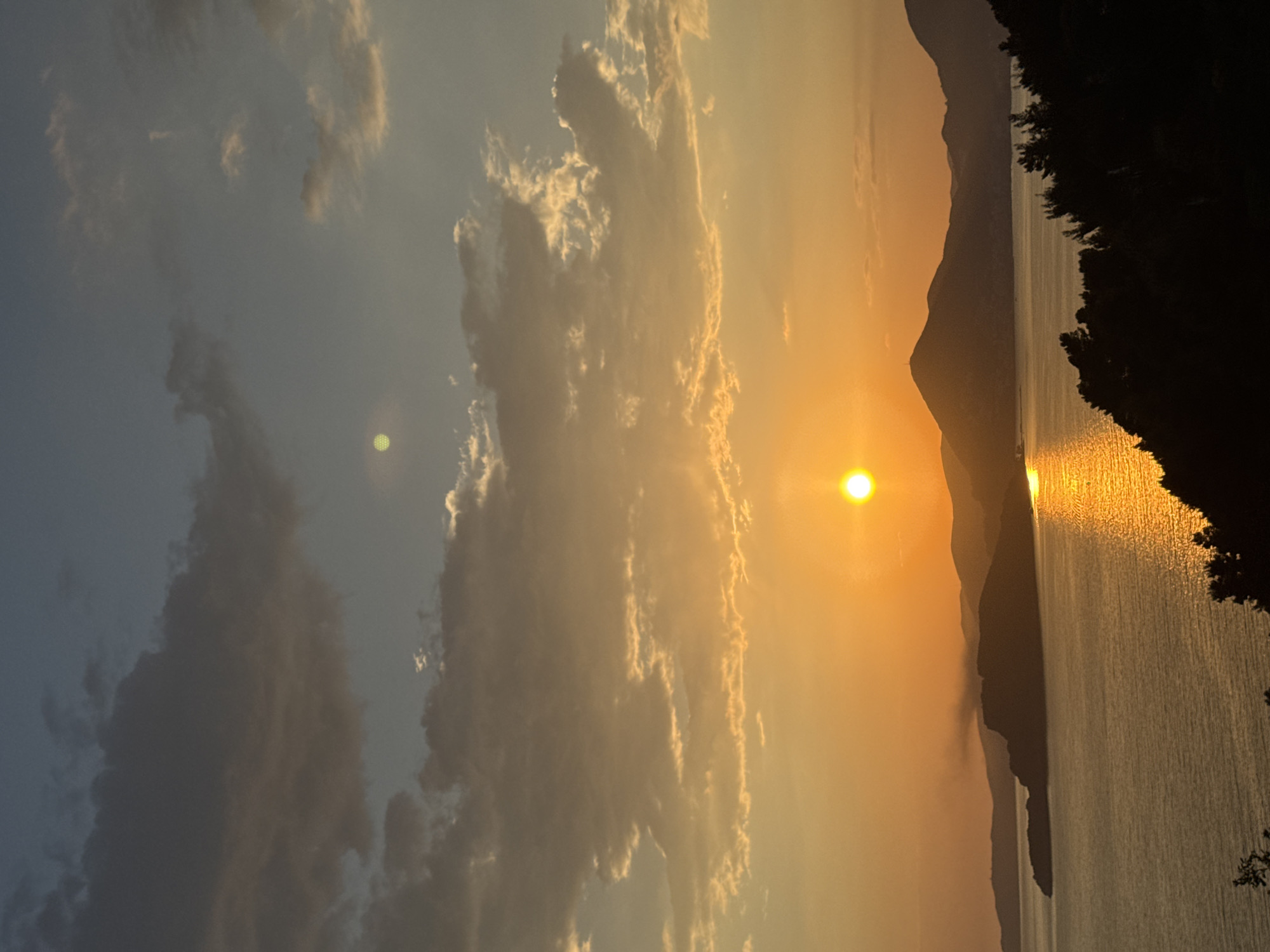 The image captures a sunset scene in Sveti Stefan, Montenegro. The sun is positioned on the horizon, casting a golden light across the sky and reflecting on the water. Mountain silhouettes are visible in the background. To the right, dark foliage of trees is present. The sky features clouds illuminated by the setting sun.