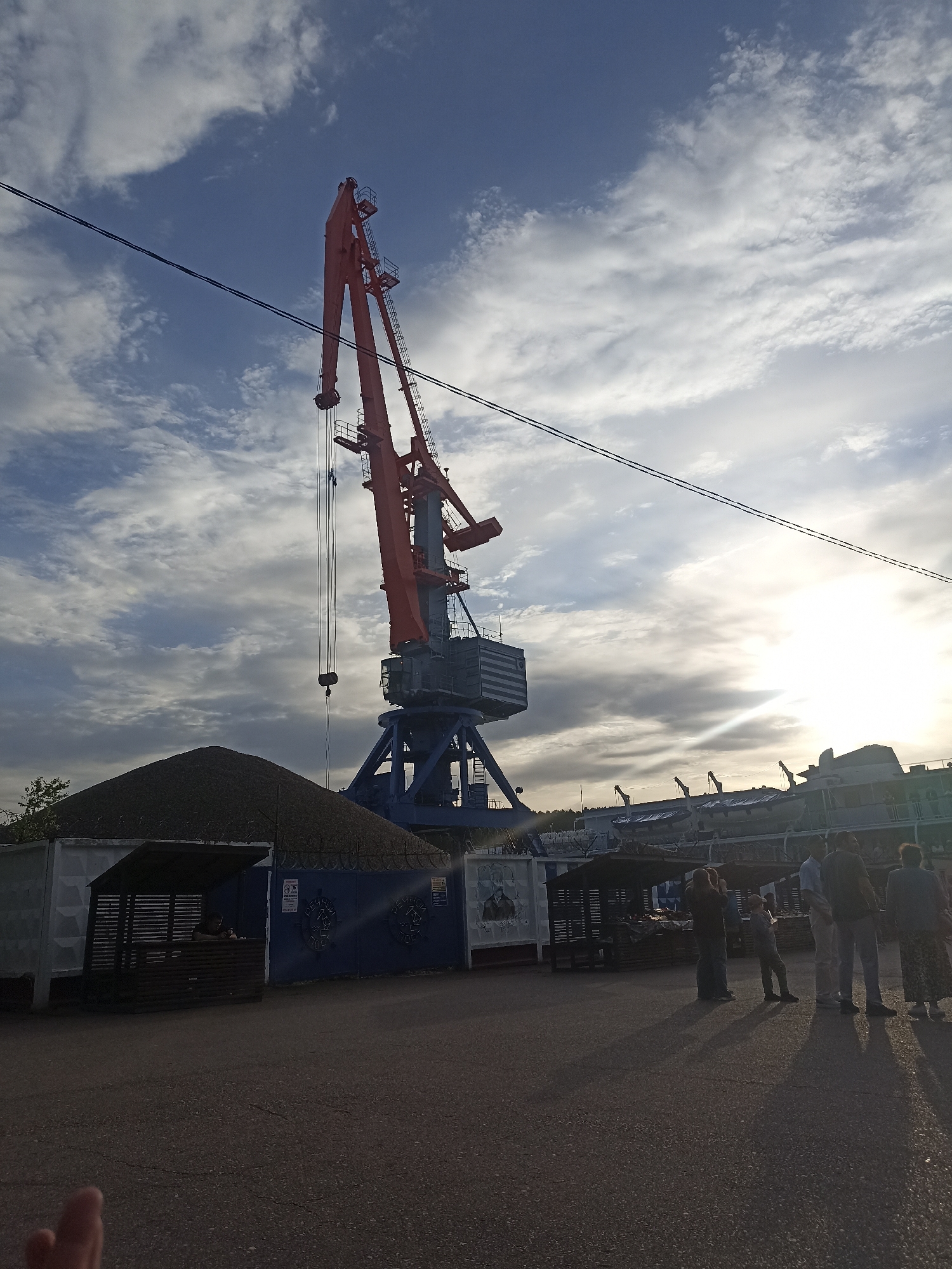The image shows a port scene in an unidentified city. A large, blue and red port crane dominates the foreground. To the left of the crane is a small, open-sided structure with a dark roof, possibly a small shop or stall. Behind the stall is a blue gate with inscriptions. To the right, a group of approximately six people are standing on a paved surface, casting long shadows. In the background, there is a river vessel docked, with several smaller cranes visible behind it. The sky is partly cloudy, with the sun obscured, giving the scene a slightly overcast appearance.