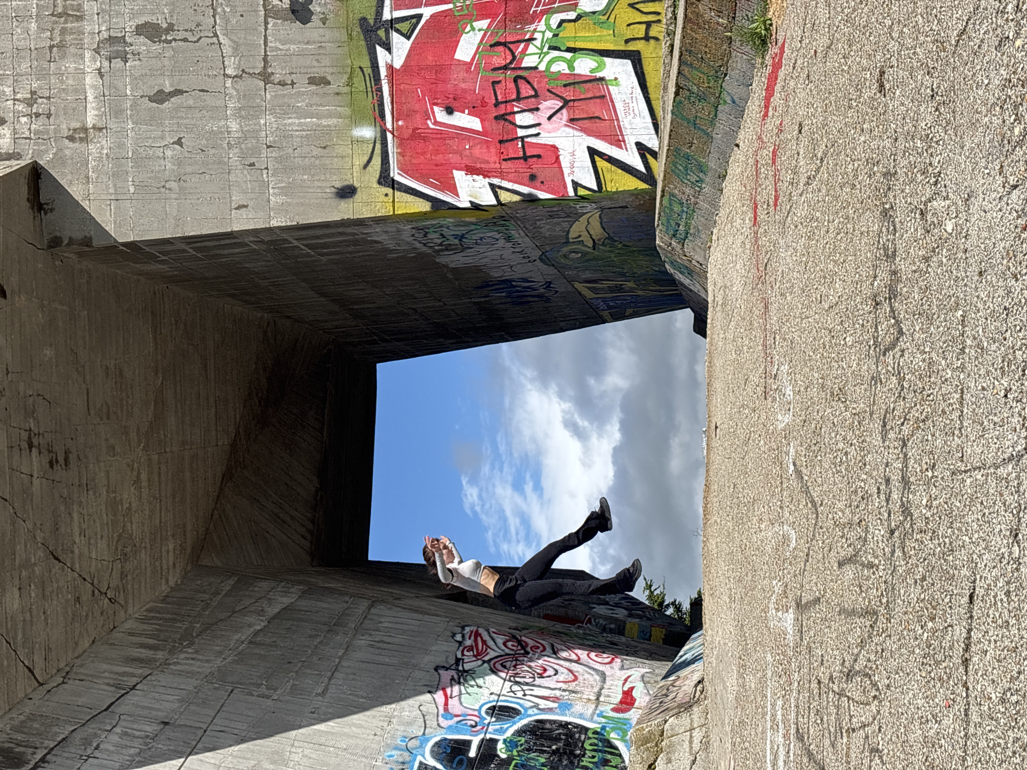 A wide shot features a woman reclining inside a concrete structure, framed against a sky with scattered clouds. The structure, likely an abandoned military site, is heavily covered in graffiti. The woman, dressed in a white top and dark pants, is positioned so that her head is slightly raised, possibly holding a cigarette, with her body extending towards the opening of the structure, where the sky is visible. The concrete walls are textured and marked with time, and the graffiti is a mix of colors and styles. The setting is reminiscent of the abandoned military sites outside Varna, Bulgaria, known for their graffiti and unique architectural shapes.