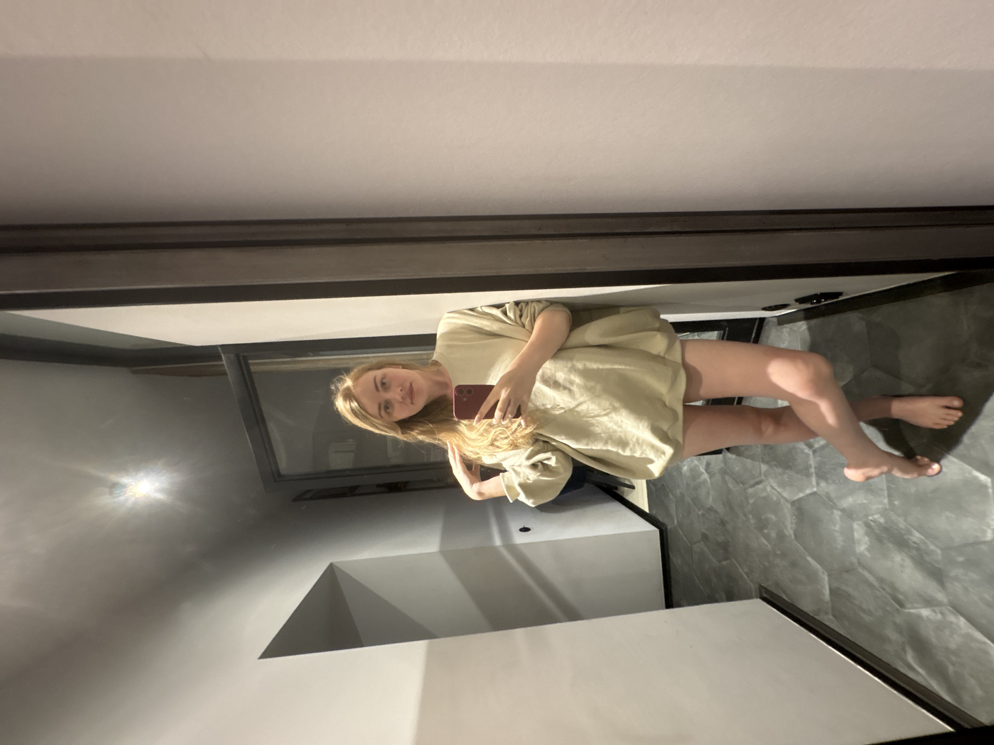 A blonde woman is standing in a bathroom with grey hexagonal tiled floors, taking a selfie in a large, dark-framed mirror. She is wearing a loose, light-colored garment. The bathroom walls are light grey, and a light fixture is visible in the mirror's reflection. No evidence suggests the location is Saint Petersburg, Russia.