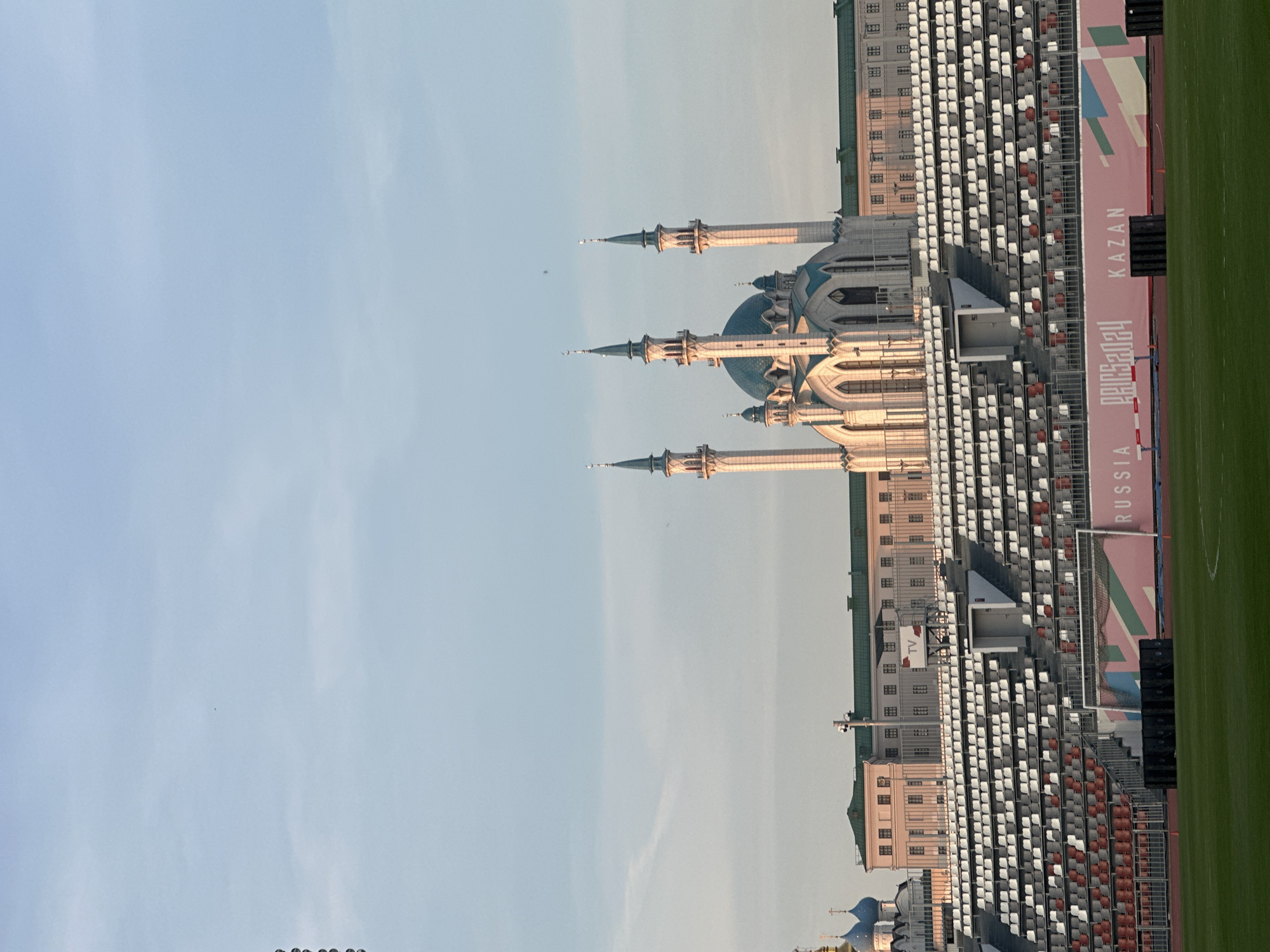 This image captures a panoramic view of iconic landmarks in Kazan, Russia, under a clear, pale blue sky, likely during late afternoon or early morning due to the soft, directional light.
The prominent subject on the left is a grand mosque, identifiable as the Kul Sharif Mosque. It features a light-colored facade, intricate arched windows, and multiple tall, slender minarets with pointed spires. Several domes crown the structure, with the largest central dome appearing blue.
To the right of the mosque, a large stadium is visible, showcasing extensive rows of empty seating. The seats are predominantly white, interspersed with sections of orange or brown seats. A vibrant green sports field or track occupies the far right of the frame. Along the edge of the field, below the stadium seating, are advertising banners or hoardings. Visible text on these banners includes "RUSSIA," "KAZAN," and "PARIS2024." A "TV" sign is also visible on a building integrated into the stadium structure.
No people are present in the scene, and the stadium appears empty, suggesting no event is currently underway. The image effectively combines the grandeur of religious architecture with modern sports infrastructure, characteristic of Kazan.