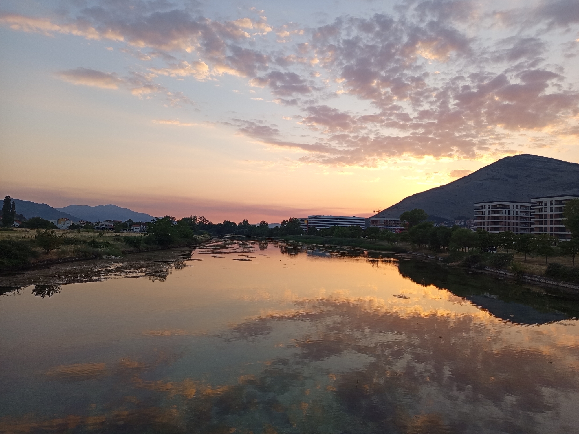 A wide-angle, landscape photograph captures a serene river scene at sunset in Trebinje, Bosnia and Herzegovina. The sky is painted with soft hues of orange, pink, and pale blue, with scattered, fluffy clouds reflecting in the calm water. In the foreground, the river stretches across the frame, its surface mirroring the dramatic sky and the surrounding landscape.
Along the left bank of the river, a collection of buildings and trees are visible, suggesting a residential or developed area. The middle ground features more foliage and structures, including a long, modern building with multiple windows, and further to the right, a series of multi-story apartment buildings with dark-trimmed windows. A steep, dark mountain rises prominently on the right side of the frame, its silhouette partially obscured by the setting sun. The overall impression is one of tranquility and natural beauty, enhanced by the warm, diffused light of the sunset.