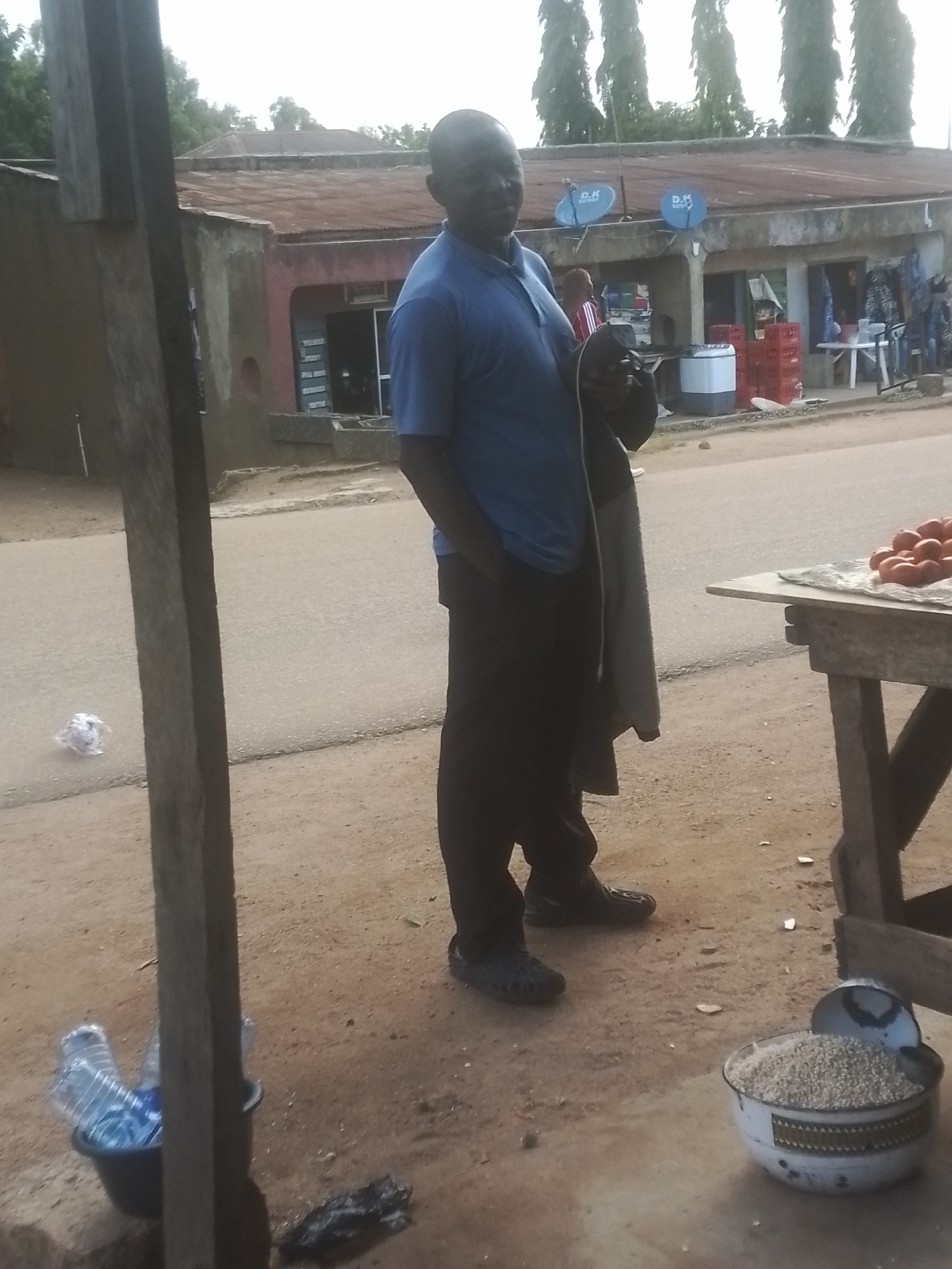 A man stands on a dirt road in front of a weathered building. He is wearing a blue polo shirt and dark pants. The building appears to be a small shop with a corrugated metal roof and several windows. There are two satellite dishes mounted on the front of the building. In front of the shop, a wooden table displays a pile of red tomatoes. To the left of the man, a black bucket contains several plastic water bottles. The ground is covered in dirt and scattered debris. The sky is overcast.