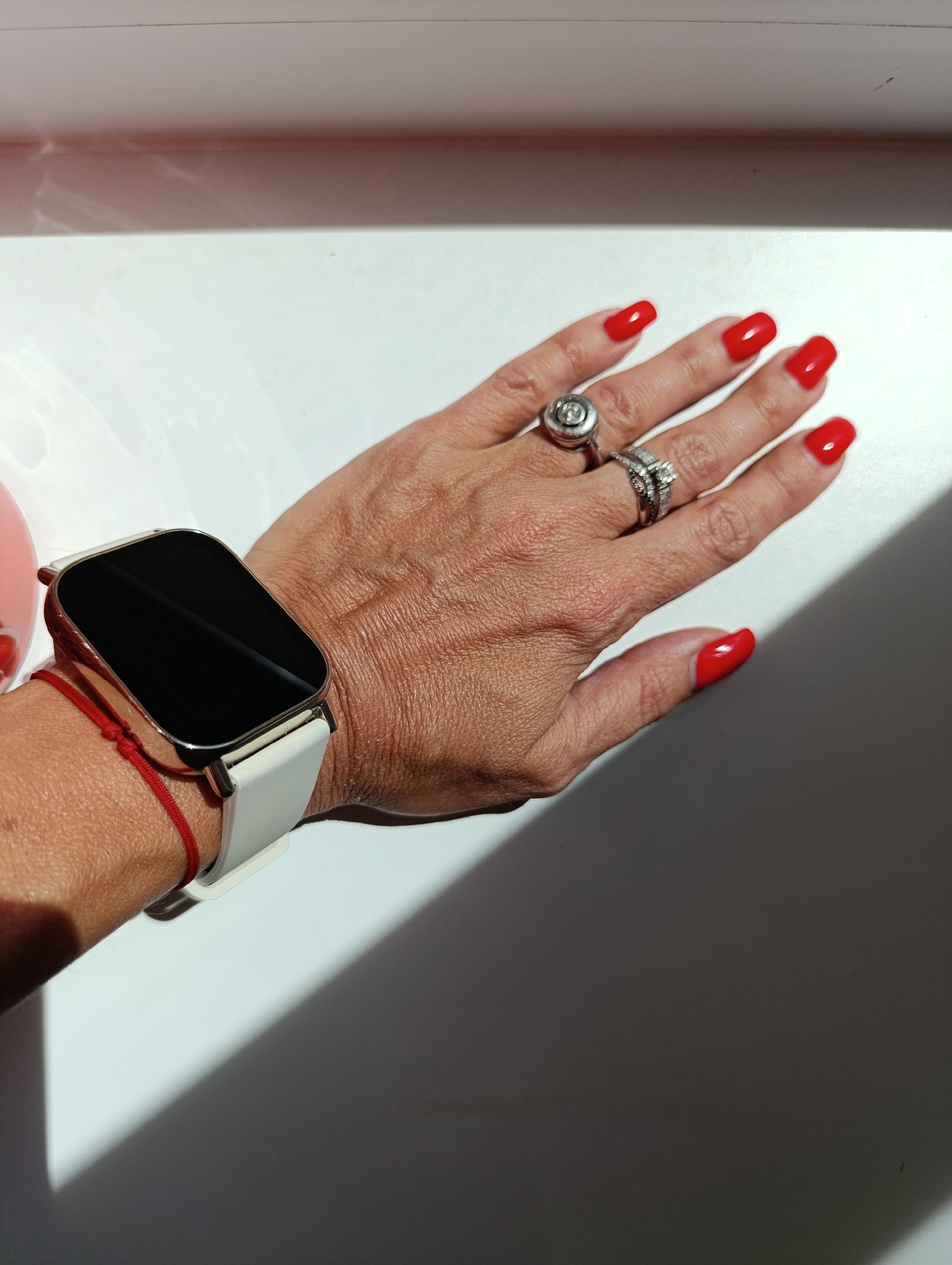 A close-up shot shows a woman's left hand with a white smartwatch on her wrist. Her nails are painted a vibrant red, and she is wearing two silver rings. One ring, on her index finger, features a circular design with a stone, while the other, on her middle finger, appears to be a band with smaller stones. A thin red string bracelet is also visible on her wrist. The background is a white, flat surface, with a harsh shadow cast across it from an unseen light source. The overall lighting suggests an indoor setting, possibly a room or a studio. The image does not contain any identifiable landmarks or features that would allow for its location to be determined in Chaikovsky, Russia, or any other city.