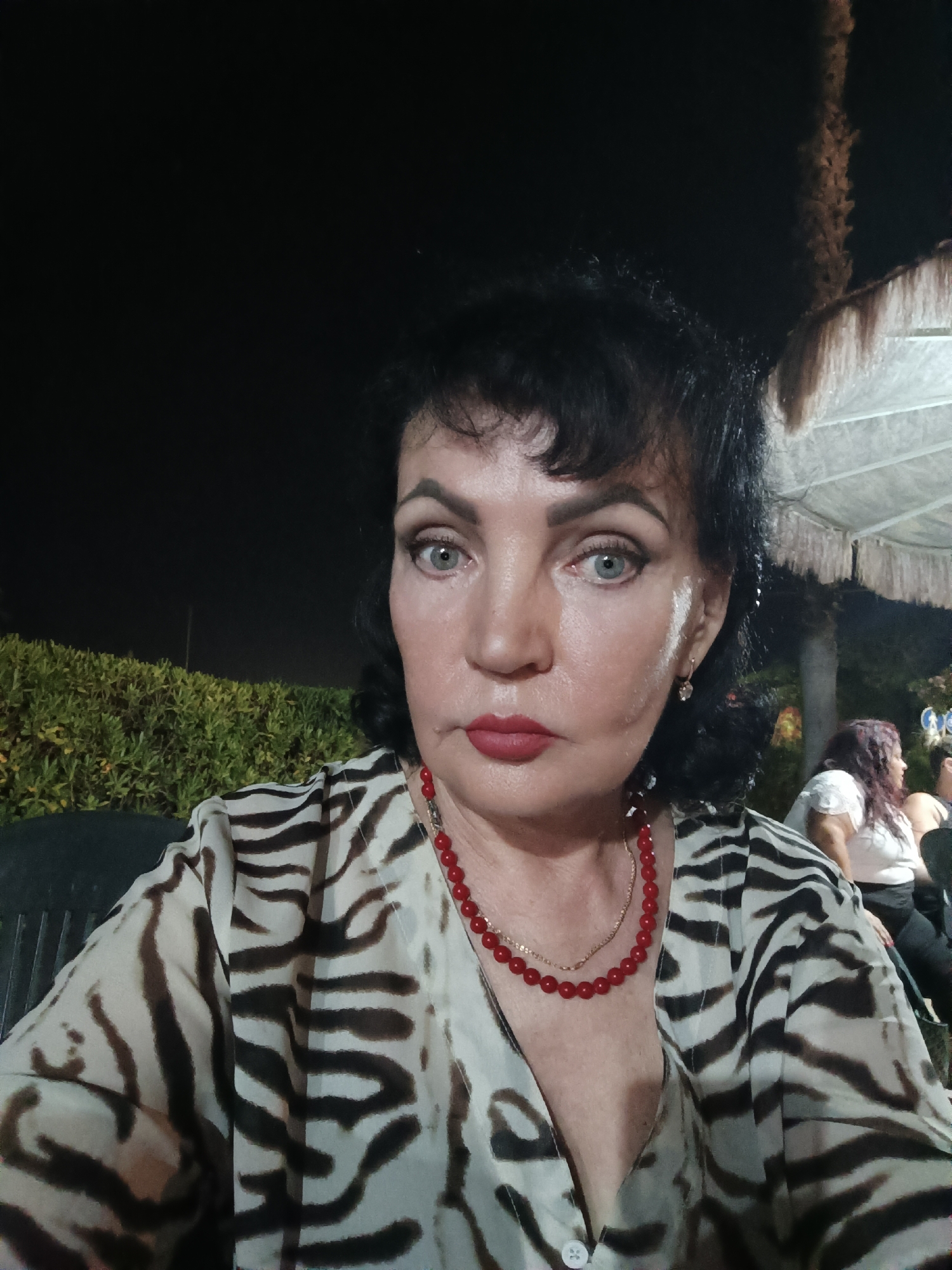 This is a medium shot selfie of a woman with dark hair and fair skin, wearing a zebra-print top and a red beaded necklace. She has on dark eye makeup and red lipstick, and she is looking directly at the camera with a serious expression. The background is dark, suggesting it is nighttime. To the left of the woman is a green hedge. To the right, a portion of a white tent with decorative trim is visible. In the background, out of focus, a woman with curly hair wearing a white top is visible. The scene appears to be outdoors, possibly at an event or gathering. The image was taken in Giulianova, Italy.