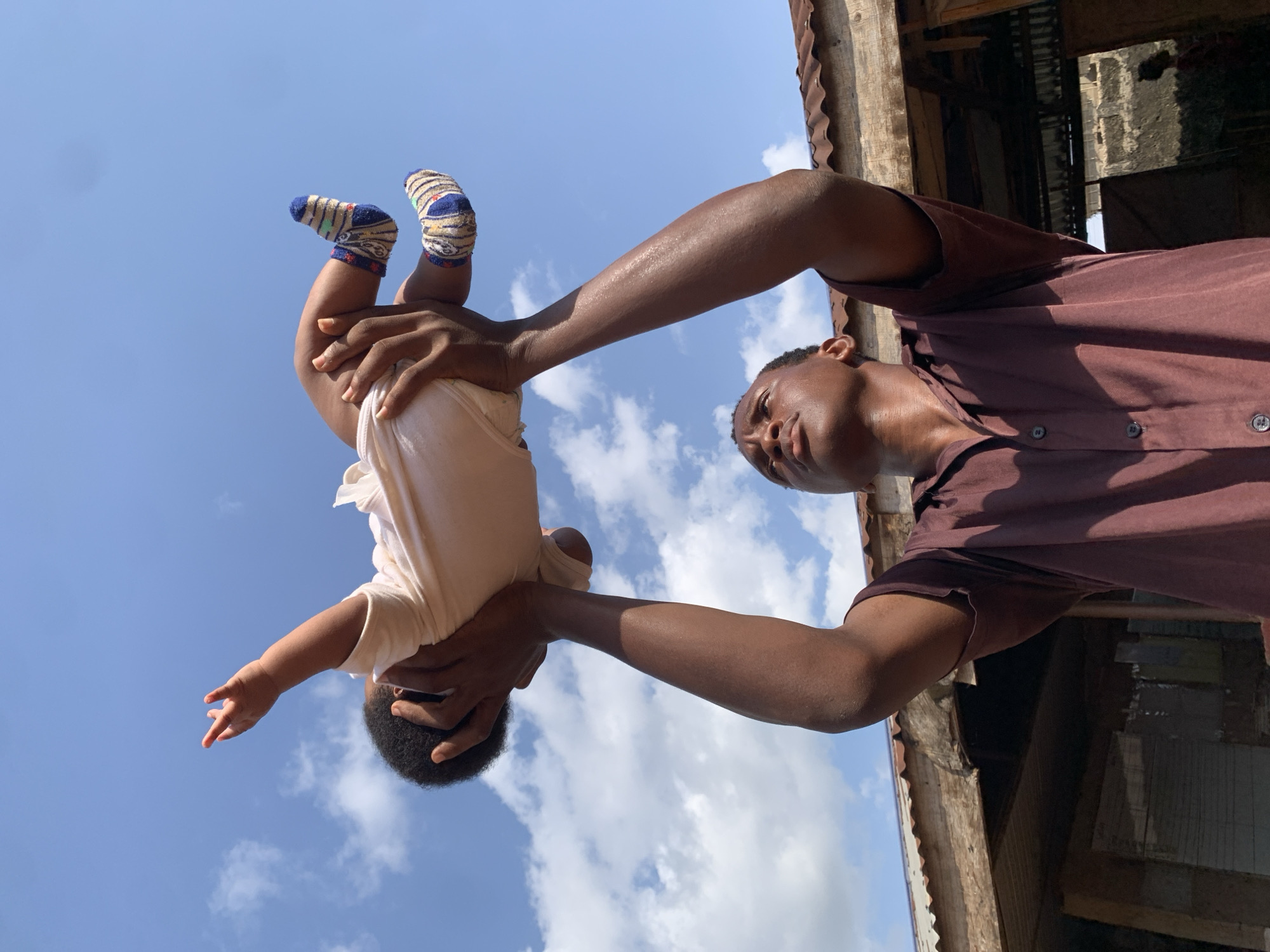 A man stands beneath a blue sky dotted with clouds, holding a baby aloft. The baby is wearing a light-colored shirt and socks with blue, white, and brown patterns. The man has dark skin and is wearing a brown collared shirt with buttons. The scene appears to be in Ukwu-Abwa, Nigeria, judging from the surrounding architecture, which includes structures with corrugated metal roofing and aged, weathered wooden beams. The orientation suggests the photo was taken from a low angle, looking up at the man and baby.