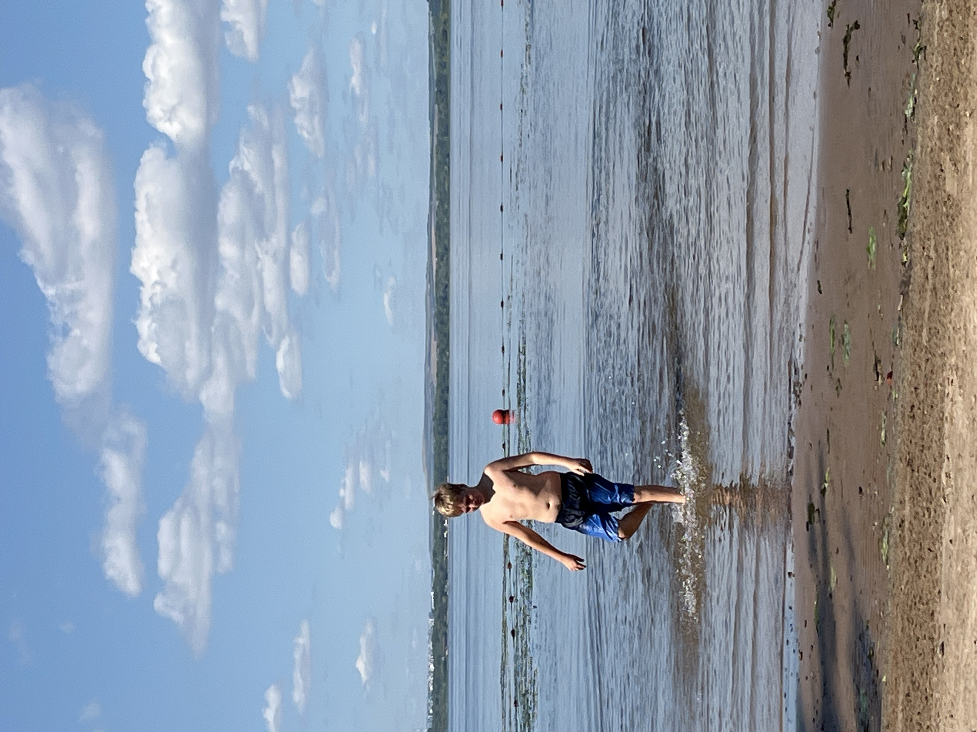 A young boy is running into the water at a beach. He is wearing blue swim trunks and is partially submerged in the shallow water, with a spray of water kicked up around his legs. The boy is shirtless, and his hair is light brown. In the background, the water stretches out to a distant tree-lined shore, with a red buoy visible on the surface of the water and a line of buoys marking a swimming area. The sky above is a bright blue with scattered white clouds. The beach itself is sandy with some vegetation. The image appears to be taken on a sunny day, likely in the afternoon. The overall scene suggests a leisurely summer day spent at the beach.