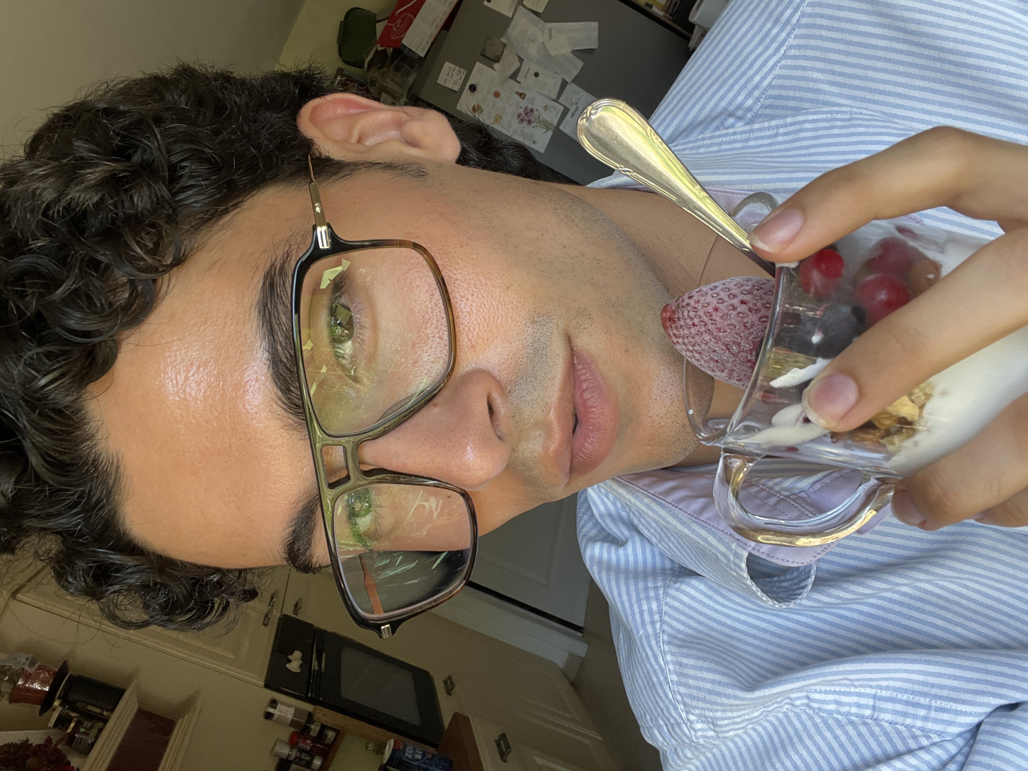 A close-up, eye-level, medium shot captures a person with dark, curly hair and light skin from the chest up, holding a clear glass cup of yogurt with berries and granola. They are wearing a light blue and white striped button-up shirt with a pink collar. The person is wearing large, rectangular eyeglasses with thick brown frames. Their right hand is holding the glass cup from the side, and their left hand is holding a gold spoon inserted into the yogurt. A single strawberry is visible on top of the yogurt. The background appears to be a kitchen, with cabinets, appliances, and shelves visible. The scene is lit by natural light, casting some shadows. The location is specified as Rotheux-Rimière, Belgium.