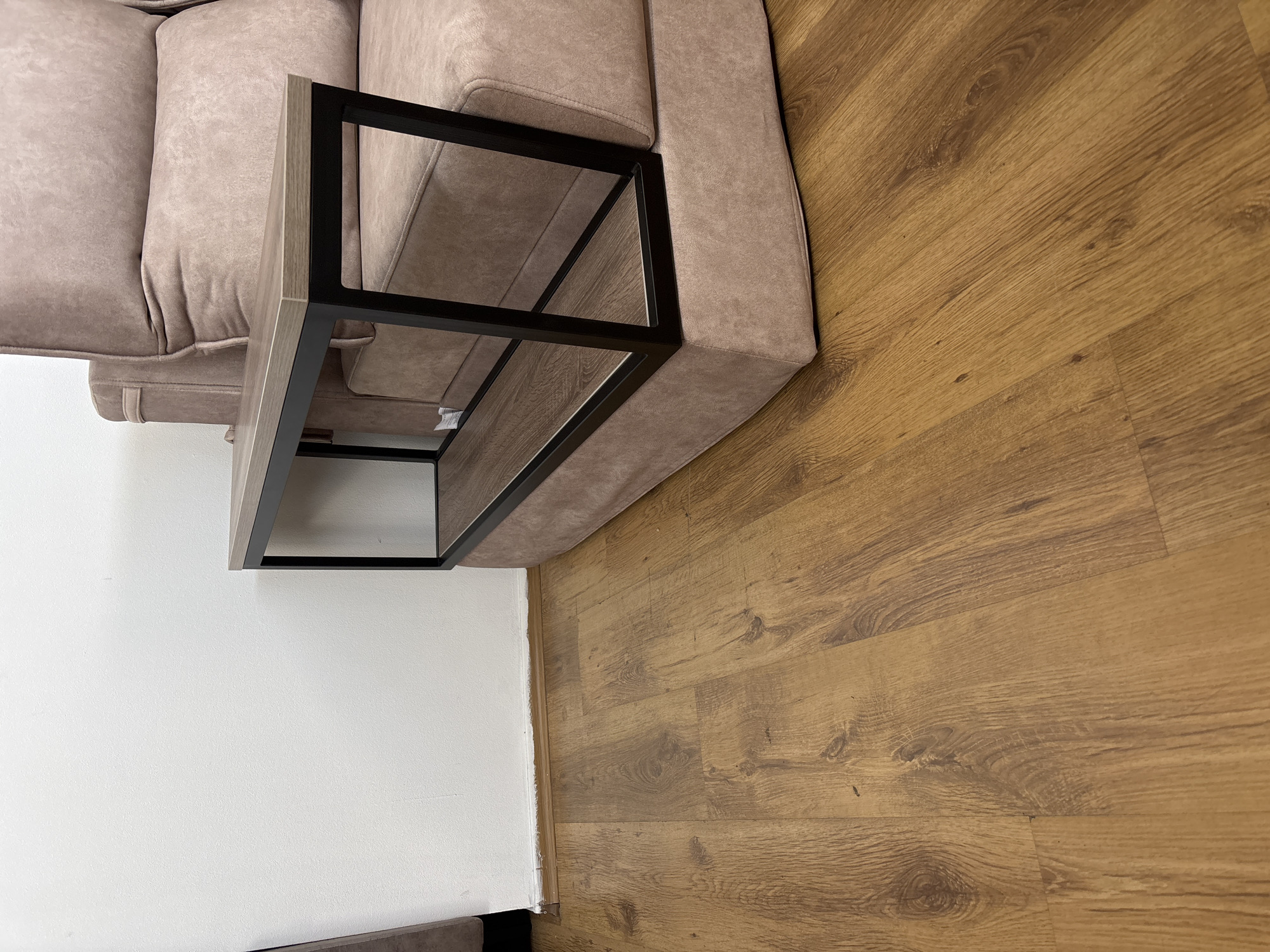 The image shows a close-up view of a modern living room setting. The primary focus is a light-colored fabric sofa and a contemporary coffee table with a black metal frame and a wood-effect top. The coffee table is positioned in front of the sofa, creating a neat arrangement. To the right of the coffee table and sofa, a warm-toned wooden floor is visible, with planks laid horizontally. The wall to the left of the sofa is painted white, providing a clean backdrop. The lighting suggests an indoor setting, likely during the daytime, though no specific time is evident. There are no people in the image. The scene is static and appears to be a staged shot showcasing the furniture and interior design. No text is visible in the image.