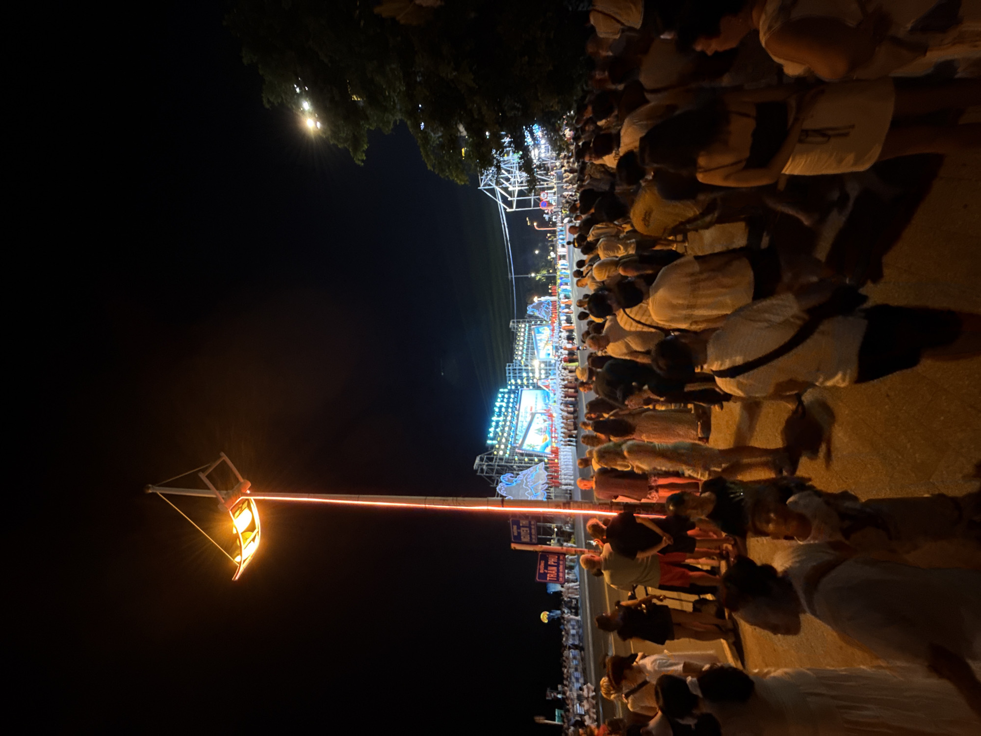 The image captures a nighttime scene on Tran Phu Street in Nha Trang, Vietnam, likely during a festival or public event. A large crowd of people, dressed in casual attire, is gathered along a street. In the background, a brightly lit stage with a large screen and illuminated decorations suggests a performance or presentation is taking place. A tall, modern lamppost with a uniquely designed illuminated fixture stands prominently in the foreground, casting a warm orange glow. Trees are visible on the right side of the image, indicating an outdoor setting. The street is paved, and a sidewalk is present. The overall atmosphere appears festive and crowded, with people observing the events unfolding.