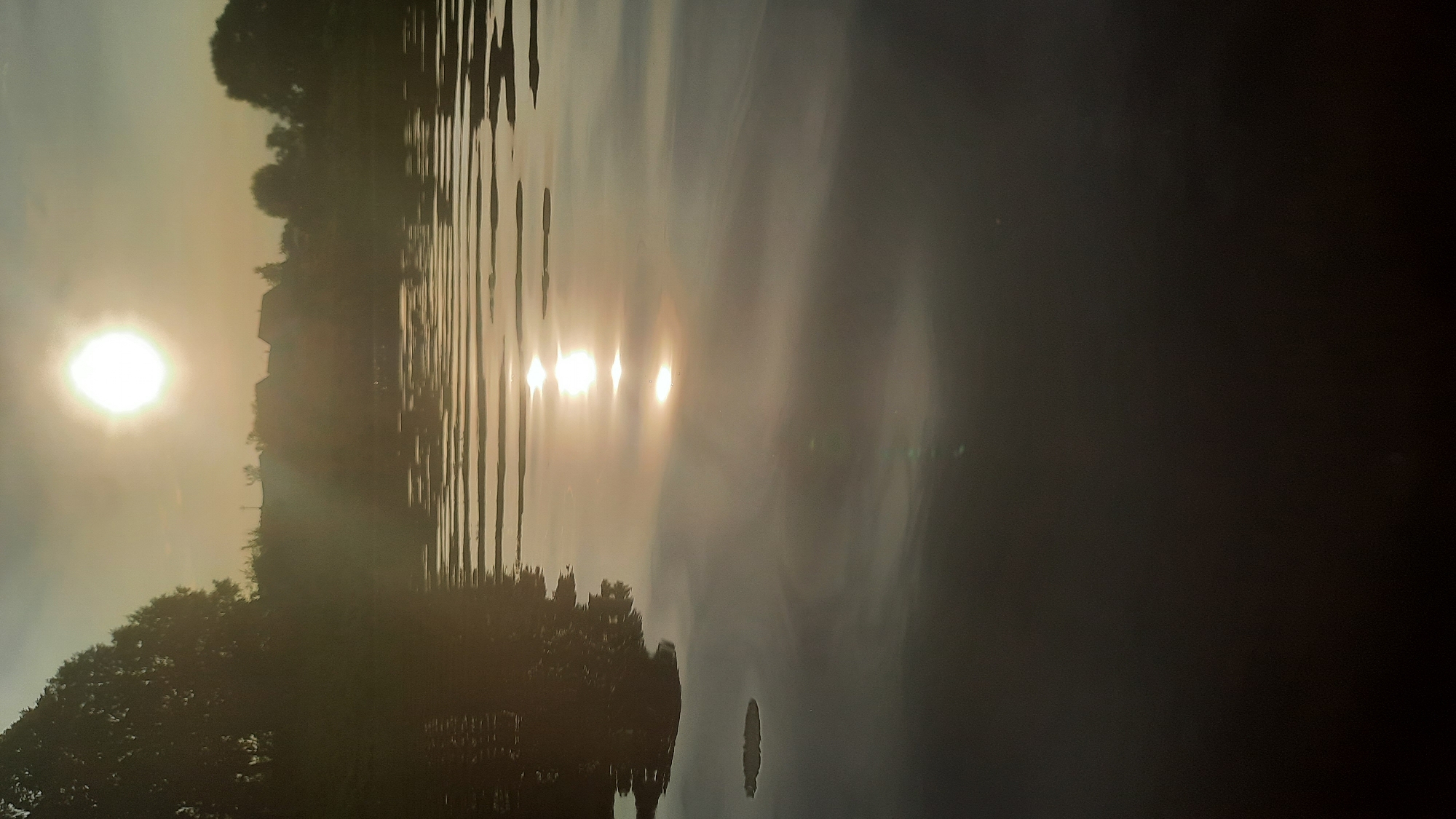 A vertical, eye-level shot captures a serene natural scene at dusk, possibly along the Dnieper River in Shklov, Belarus. The sun, a bright, hazy orb, sits high in the sky, casting a warm, golden glow. Its reflection shimmers on the rippling surface of the water, creating streaks of light. Trees line the distant shore, their silhouettes softened by the low light. A few structures, likely houses, are visible among the trees, suggesting a rural or semi-rural setting. The foreground of the water is dark and reflective, with subtle ripples creating patterns. The overall atmosphere is peaceful and tranquil, evoking a sense of calm as day transitions to night.