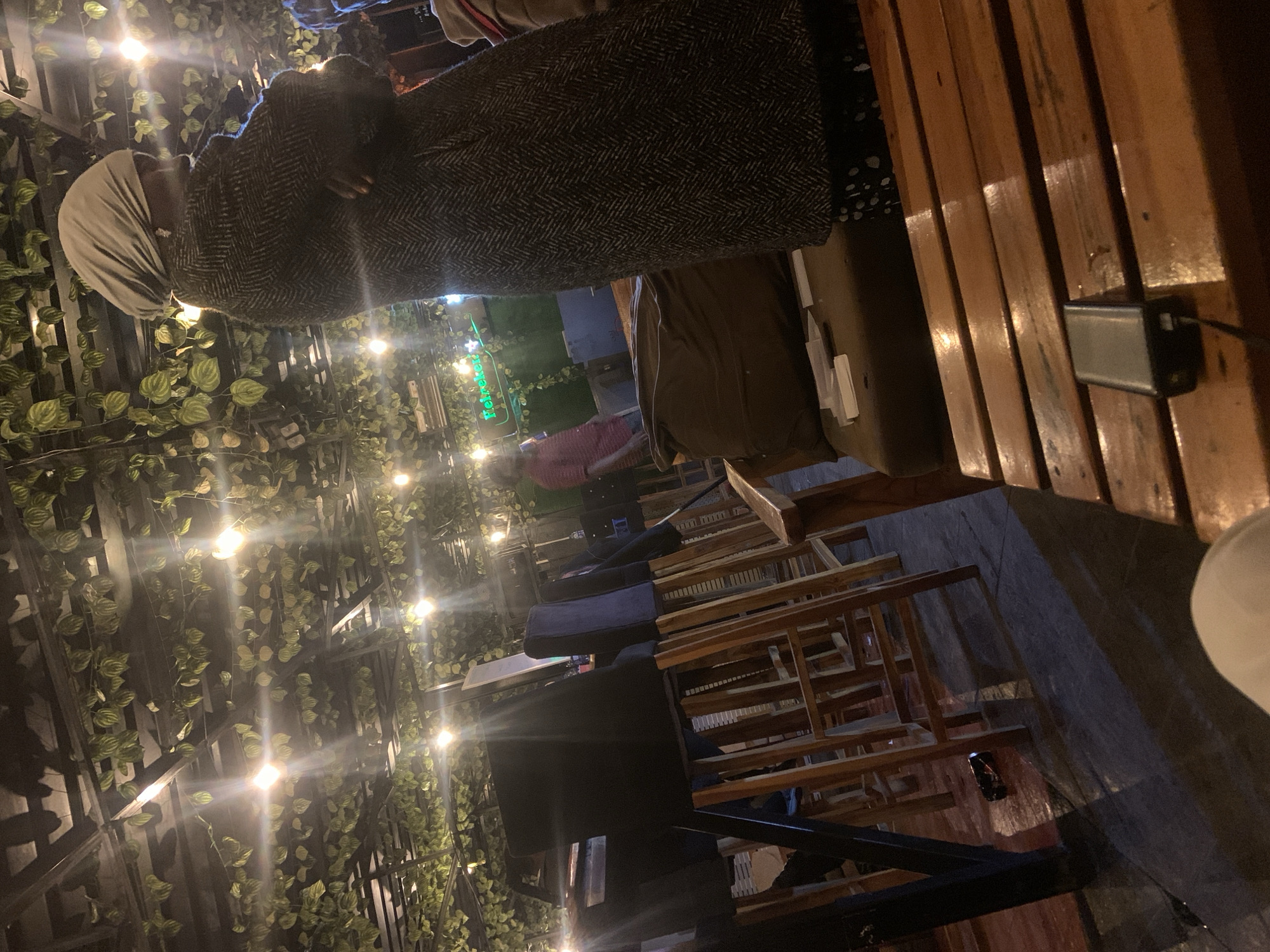 The image depicts an outdoor seating area, possibly a restaurant or bar, decorated with artificial greenery and string lights along the ceiling. In the foreground, a person wearing a grey headscarf and a patterned coat is standing with arms crossed. A wooden table with a power bank on it is also visible in the foreground. In the background, there are several wooden bar stools and tables, and a person in a red shirt can be seen standing near a neon sign that reads "Heineken." The overall ambiance suggests a lively evening setting in Gura Top, Nigeria.