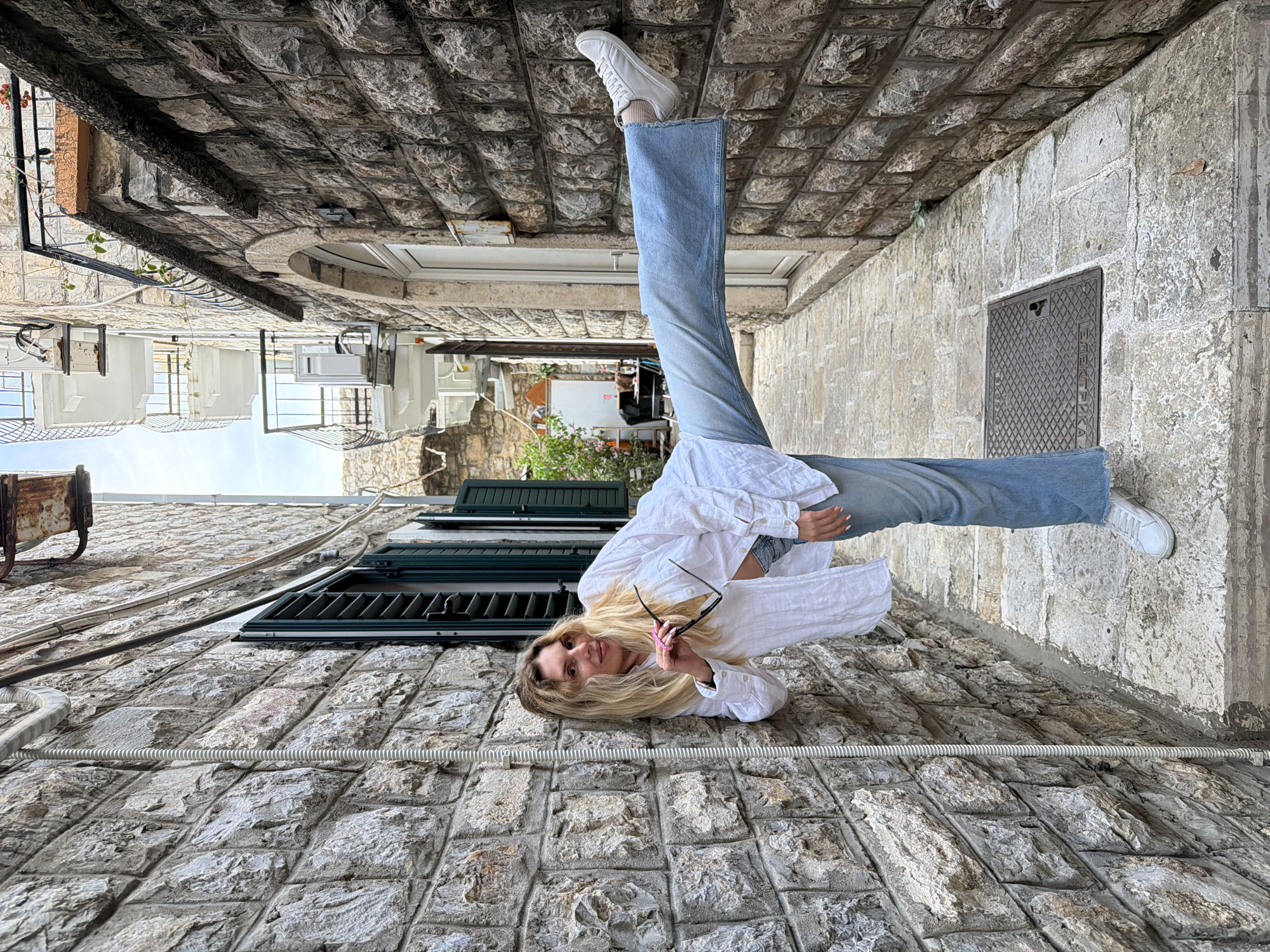 The image displays a young woman striking a dynamic pose in a narrow, stone-paved alleyway. She is wearing a white button-down shirt, blue wide-leg jeans, and white sneakers. Her body is contorted: she leans against one rough-hewn stone wall with her back, her right leg extended forward with a white sneaker, and her left leg bent and raised, resting against the opposite stone wall. She holds dark-framed sunglasses in her right hand, which has pink-painted fingernails, and looks directly at the camera with a neutral to slightly smiling expression. Her long blonde hair frames her face.
The scene is an ancient-looking alleyway with tall walls constructed from large, irregular stone blocks. The ground is also paved with similar stone. In the background, beyond the woman, other buildings with balconies, air conditioning units, and glimpses of sky and foliage are visible, suggesting an urban environment. One wall features dark green louvered window shutters. Various cables and conduits run along the walls and across the ground.
The photo is taken during the daytime under bright, clear skies. A notable detail on the ground is a cast-iron manhole cover with a diamond pattern, bearing the Cyrillic text "ЕЛЕКТРИКА" (ELEKTRIKA), which means "Electricity". The architecture and the presence of Cyrillic text strongly suggest a location in a Slavic-speaking region, consistent with a Mediterranean old town.