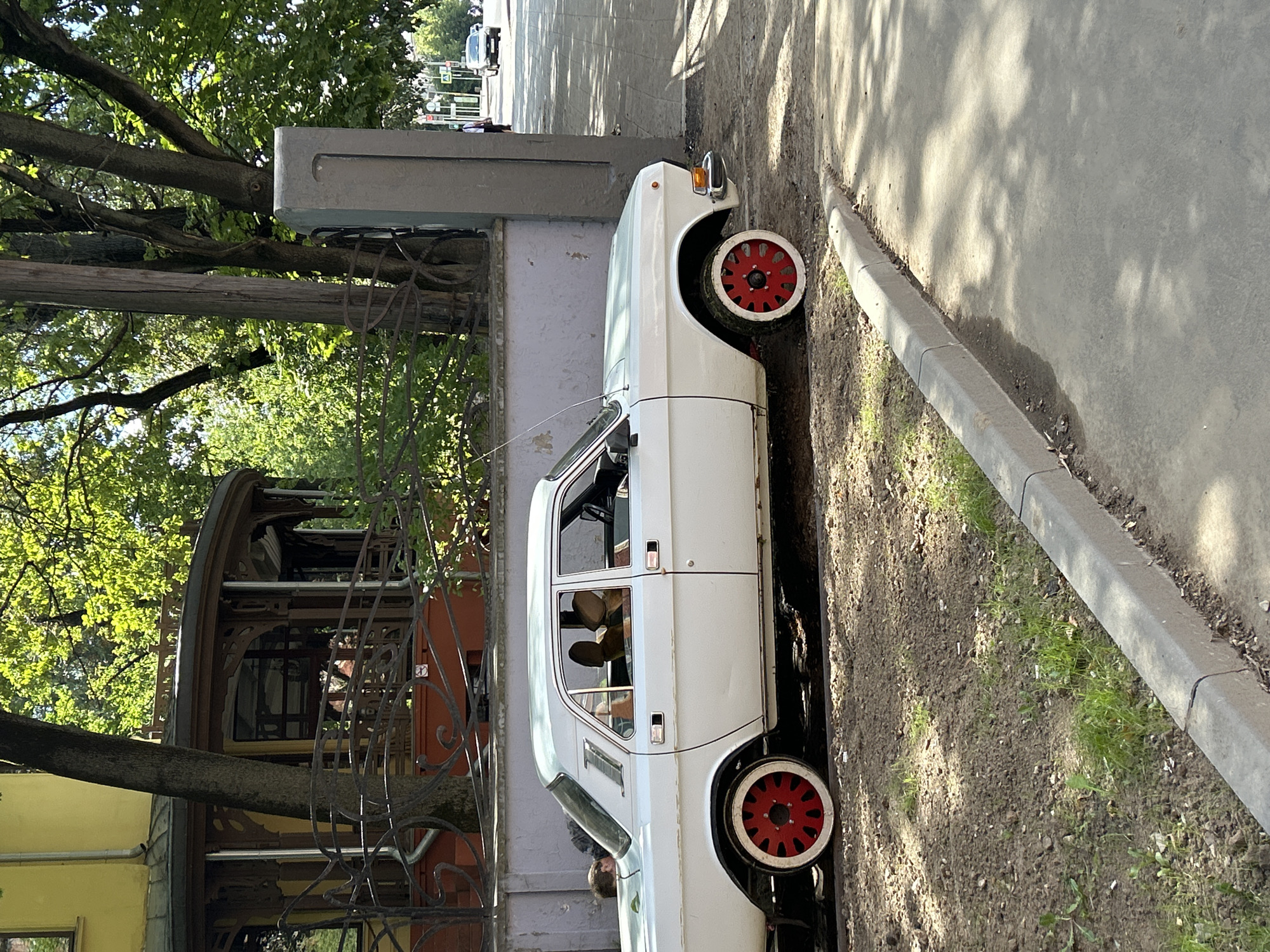 A white, vintage four-door sedan is parked on a dirt and gravel shoulder next to a paved sidewalk in a city. The car has distinct red wheels with white tires and appears to be modified. The interior of the car shows two headrests in the front and what appear to be fabric seats. A person's head and shoulders are visible through the rear passenger window, and another person is partially visible leaning into the trunk of the car. In the background, there is a building with a yellow facade and a decorative, multi-level porch or balcony. Trees with lush green foliage and a stone pillar are also visible. A street with traffic and pedestrians is in the far background. The overall scene suggests an urban environment on a sunny day.