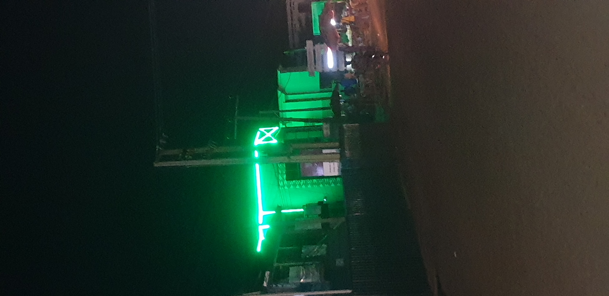 The image depicts a nighttime street scene in Sabon Tasha, Nigeria. The foreground shows a dark, unpaved or asphalt road surface. In the middle ground, a building is prominently illuminated by a bright green neon light display. This display features a horizontal green bar with a vertical segment forming a stylized 'T' shape on the left, and a distinct 'X' shape to its right. The intense green light bathes the building facade and surrounding areas. A weathered utility pole with attached wires stands in front of the illuminated structure, slightly right of center. To the right side of the image, along the roadside, several indistinct individuals are present, gathered near at least two market-style umbrellas or canopies, one appearing reddish-brown. A motorcycle or similar vehicle with an illuminated headlight is visible in the mid-right, facing away from the viewer. The sky above is dark, indicating night, with very low ambient light making details outside the neon's glow largely indistinguishable.