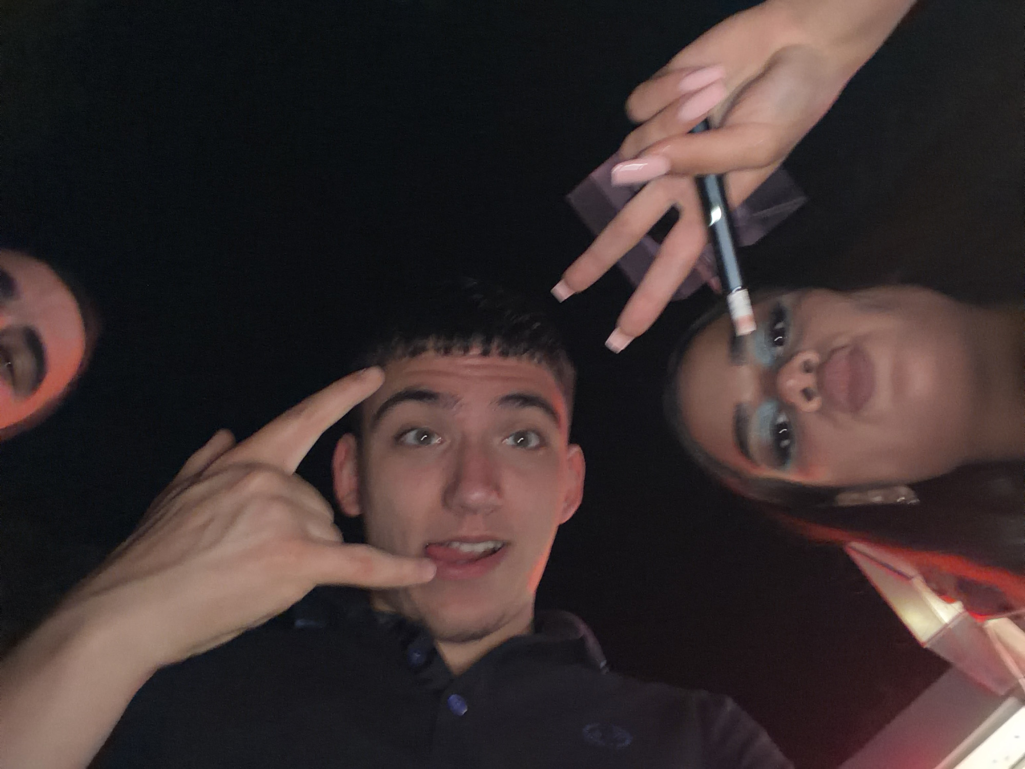 A low-angle selfie captures three individuals in a dimly lit setting, suggestive of nighttime or an indoor venue in Varna, Bulgaria. In the center, a young man with short dark hair and light eyes looks up at the camera with a playful expression, his tongue slightly out. He forms a "shaka" or "call me" hand gesture with his left hand and wears a dark polo shirt.
To the right, a young woman's face is seen upside down from the low perspective. She has noticeable light blue eyeshadow and appears to be applying makeup with a brush, held by a hand with long, light-colored acrylic nails. Her lips are slightly parted. On the far left, a portion of a third person's face, including an eye, is visible, also looking upwards. The background is predominantly dark, with a glimpse of a red and white illuminated structure or object in the bottom right corner of the frame. The overall atmosphere is casual and social. There is no visible text in the image.