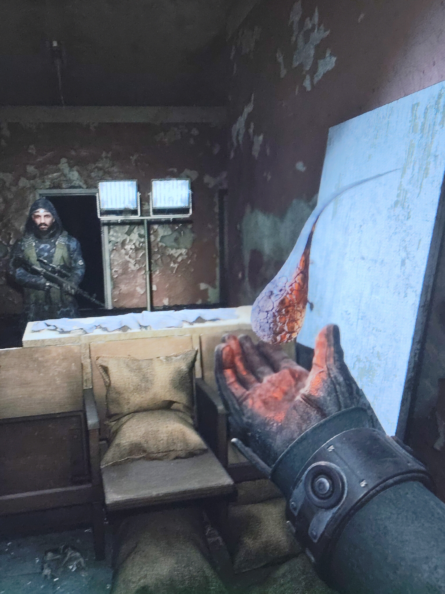 The scene is an interior room in a state of disrepair. Paint is peeling off the walls, which are colored in shades of brown and red. The floor is covered in debris. The room contains an improvised seating area with wooden chairs and cushions made of rough fabric. In the foreground, a gloved hand is outstretched, holding a pulsating, orange-tinged object. In the background, standing in a doorway with two window-like grilles above it, is a man armed with a rifle. He is wearing camouflage clothing and a hood. The room is dimly lit. The city and specific location are Unknown.