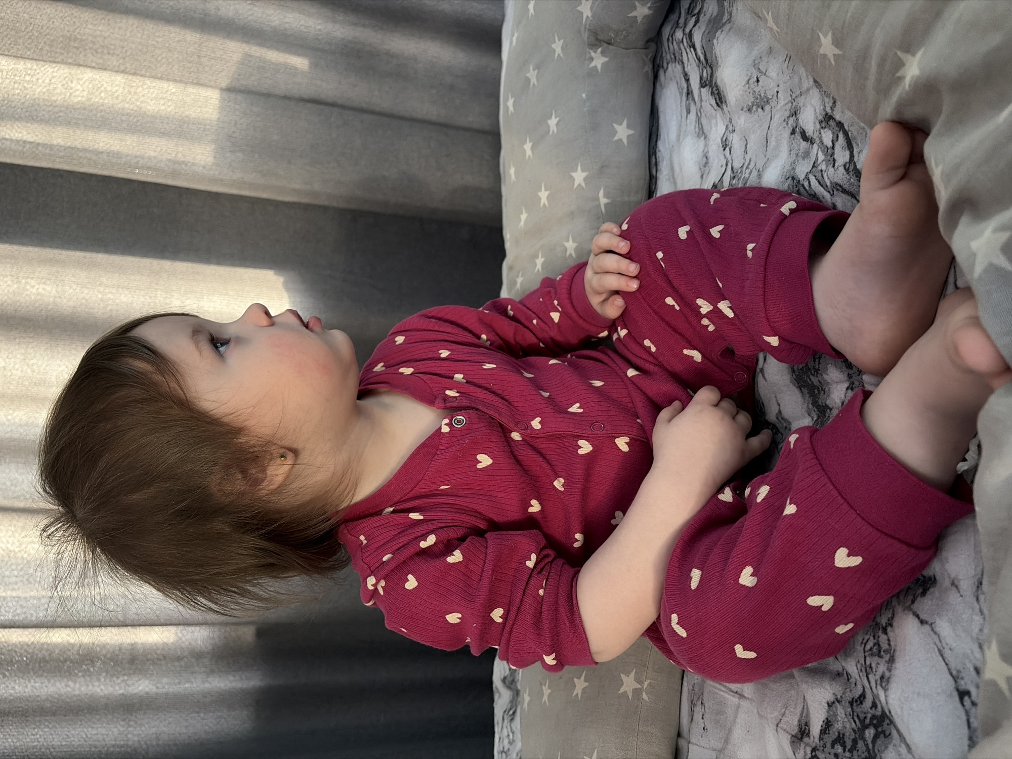A young child is sitting on a bed indoors in Chaikovsky, Russia. The child, with brown, disheveled hair and blue eyes, wears a magenta ribbed one-piece pajama suit adorned with small, light-colored heart patterns. A single gold stud earring is present in the child's visible left ear. The child is positioned with bent legs, bare feet, and hands resting on the lap, looking towards the right of the frame. The bedding beneath the child consists of a dark grey and white marbled patterned sheet. Behind and to the child's right, a light grey pillow with white star patterns is visible. In the background, light grey, textured curtains are illuminated by natural light entering from the left, casting subtle shadows.