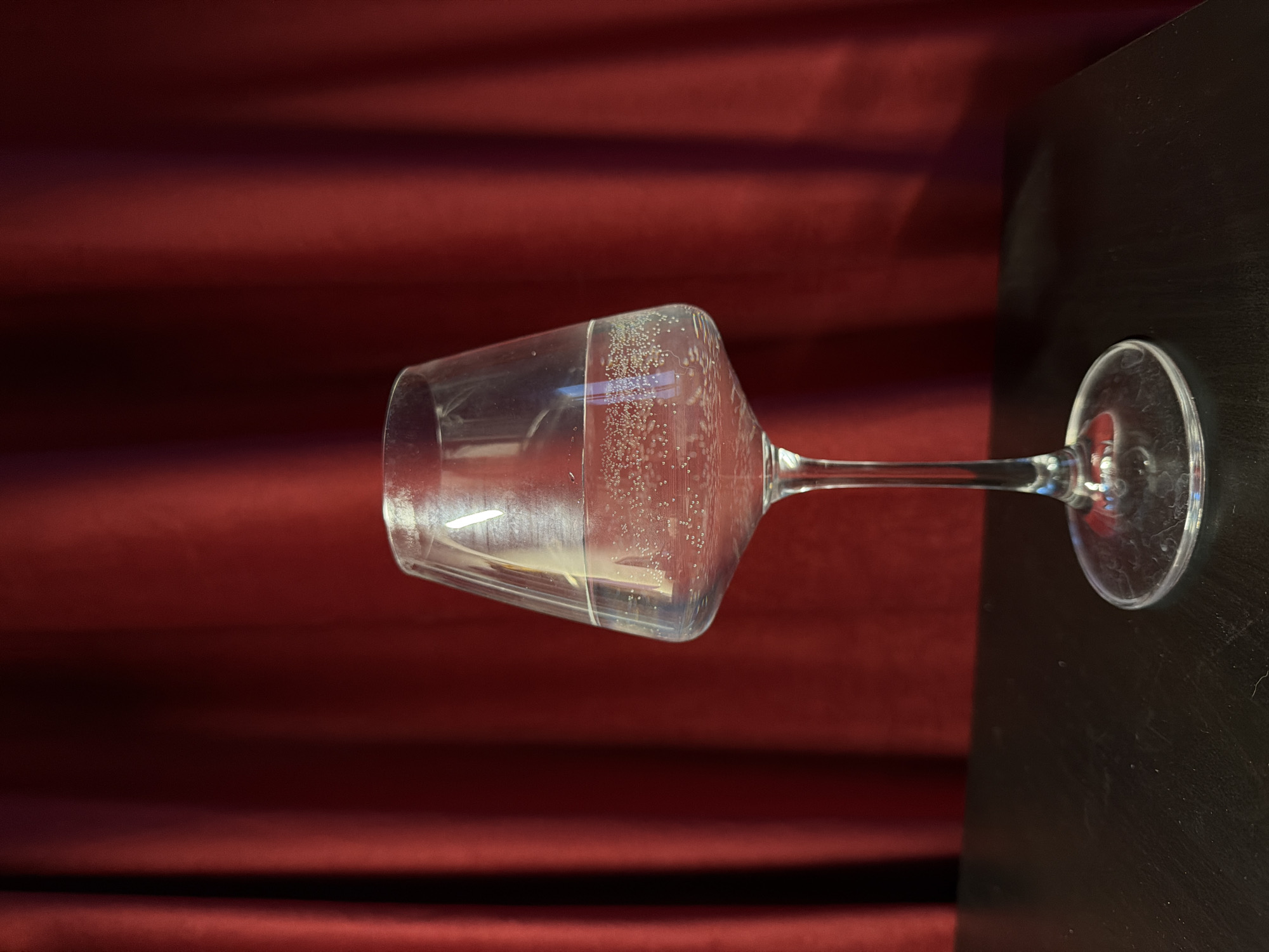 A wine glass containing a clear liquid rests on a dark, possibly wooden surface. The stem and base of the glass are on the right side of the image. A red, slightly textured background is visible behind the glass.