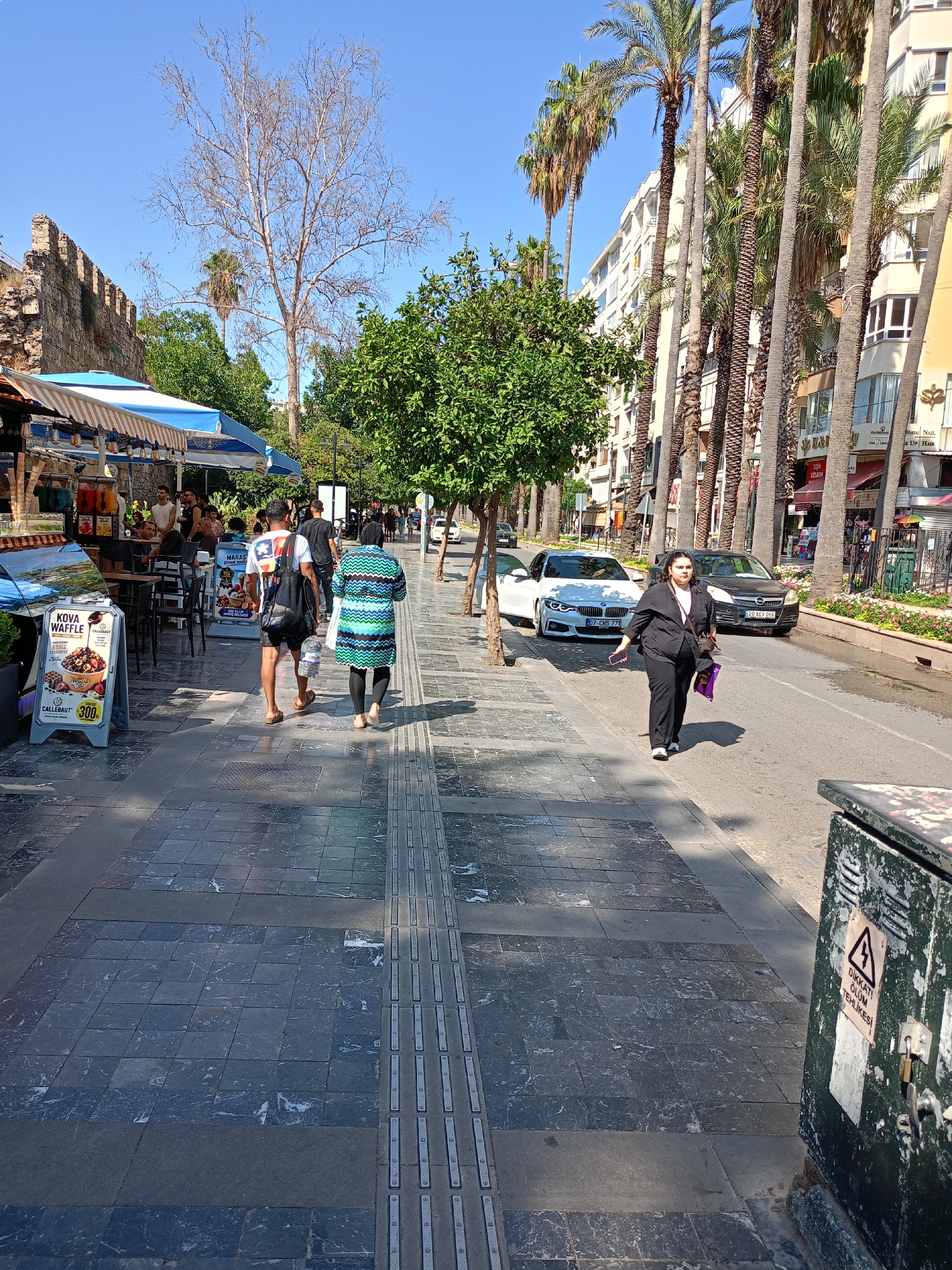This image captures a street scene in Antalya, Turkey, likely in the historic Kaleiçi district, characterized by its narrow streets, ancient walls, and palm trees. In the foreground, a woman in a black suit walks on the right side of the road, carrying a purple bag and a phone. She is walking away from the camera. On the left, a tiled sidewalk is busy with pedestrians. A man in a white t-shirt and shorts walks away from the camera, holding a water bottle. Beside him, a woman in a green and blue patterned coat also walks away from the camera. Shops with awnings line the left side of the street, displaying goods and menus. Behind the shops, the ancient stone walls of Antalya are visible. Palm trees and other greenery line the street, with modern buildings in the background. A white BMW and a dark sedan are parked on the right side of the road. A green electrical box with caution signs is visible in the lower right corner. The sky is clear and blue, indicating a sunny day.