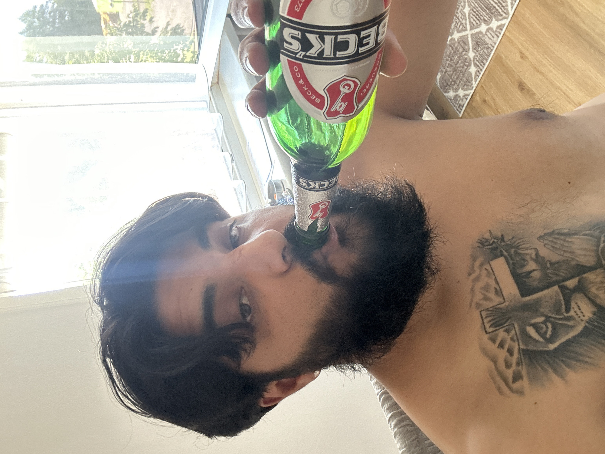 A man with dark hair and a beard is drinking from a green glass bottle with a Beck's beer label. He has a tattoo on his chest of a lion and praying hands with a cross and a crown of thorns. He is indoors, in front of a window with sheer white curtains. The environment appears to be a home with wooden flooring and a patterned rug visible. There is no indication of the city of Mainburg, Germany in the image.
