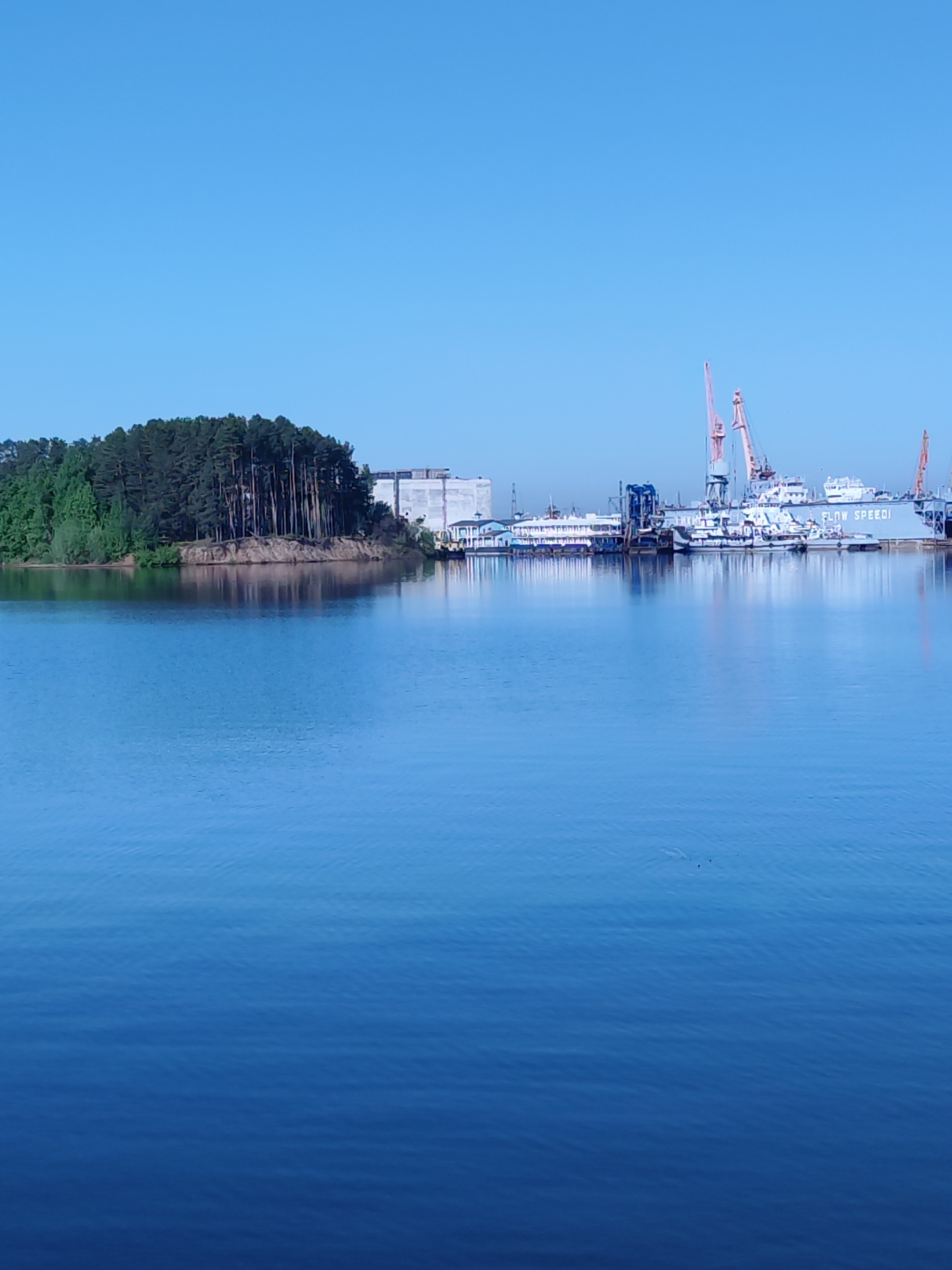 The image presents a water scene with land features and industrial activity in the background. In the foreground, a body of water, reflecting the sky and the distant objects, dominates the view.
To the left, a landmass covered in trees provides a natural contrast to the industrial elements on the right. The trees are a mix of evergreen and deciduous varieties.
In the background, on the right side of the image, there is a shipyard or dockyard with cranes and vessels. The ship "FLOW SPEED I" is visible. The sky is clear.