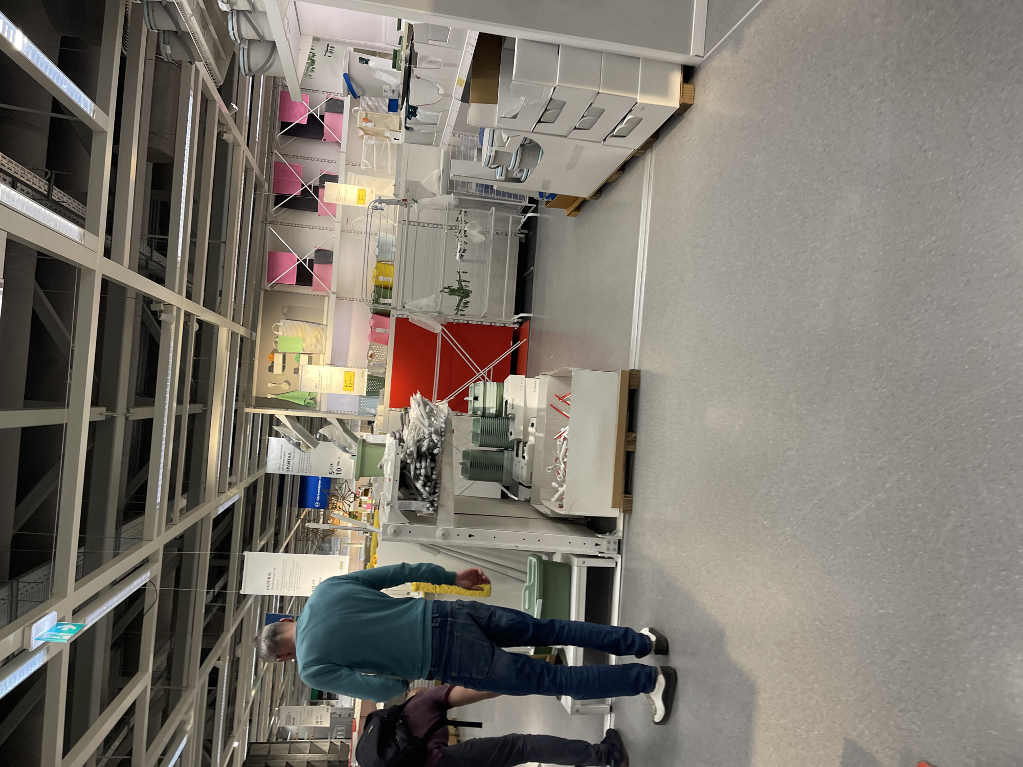The image shows the interior of an IKEA store, likely in Sofia, Bulgaria given the context. The store displays its merchandise on tall shelves and in organized displays. The building structure includes high ceilings with visible lighting and metal beams. Two individuals are visible. One, wearing a blue shirt and jeans, appears to be looking at the merchandise. The second is wearing a dark shirt and pants and a backpack.