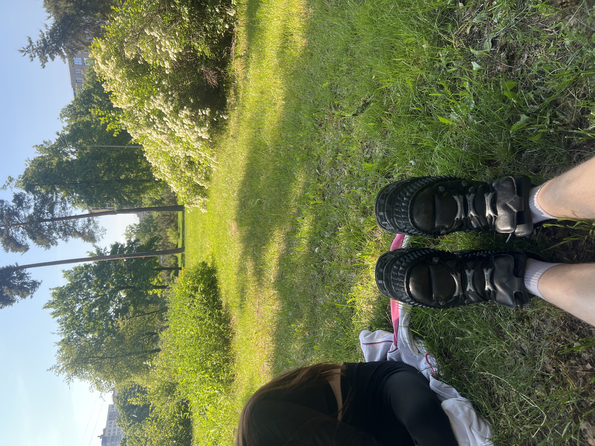 The image, captured from a first-person perspective, depicts a person resting in a sunny park. In the immediate foreground, the person's lower legs and feet are visible, clad in white socks and robust black rollerblades featuring a protective, ridged design. A white garment with a pink stripe and a black long-sleeved top are partially seen, indicating the person is seated on the grass.
The scene unfolds in a vibrant green park environment. A wide expanse of well-maintained grass fills the middle ground, brightly lit by daylight, with distinct shadows suggesting either morning or late afternoon. Along the left side of the grass, a large bush is covered in abundant clusters of small white flowers, indicative of spring or early summer.
Further into the background, numerous trees of varying heights and types, including tall conifers and leafy deciduous trees, line the park. Above the treetops, a clear blue sky is visible. Beyond the greenery, multi-story urban buildings can be discerned, confirming an urban park setting in Saint Petersburg, Russia. The image conveys a moment of relaxation and respite amidst nature. No distinct text is clearly legible on the rollerblades or other objects.