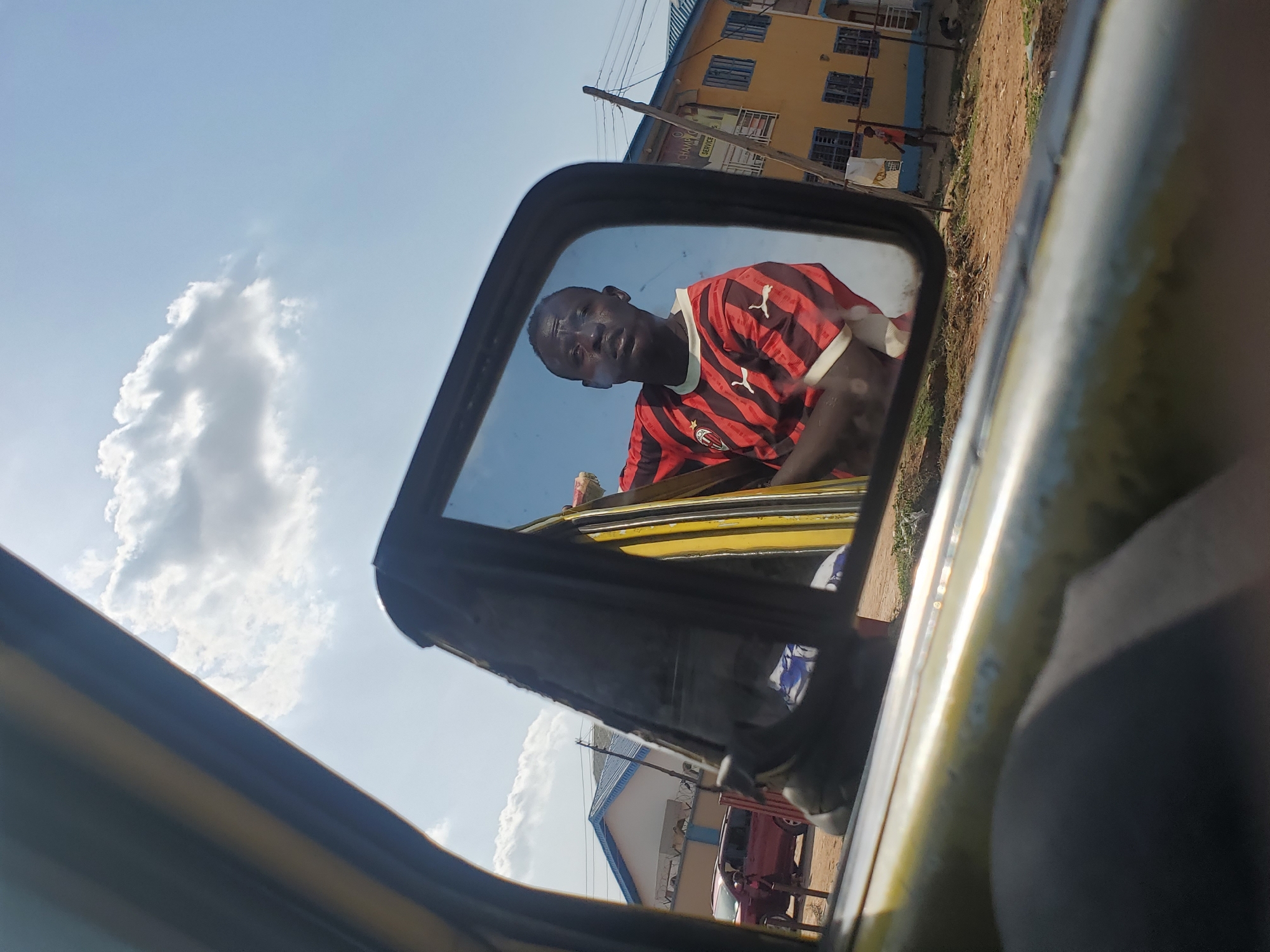 A man is reflected in a car's side mirror. He is wearing a red and black striped shirt with a white Puma logo. The car's frame is yellow. In the background, outside the car, a multi-story yellow building with blue window frames and a red car are visible, suggesting an urban setting. White clouds are visible in the sky. This image was taken in Yelwa, Nigeria.