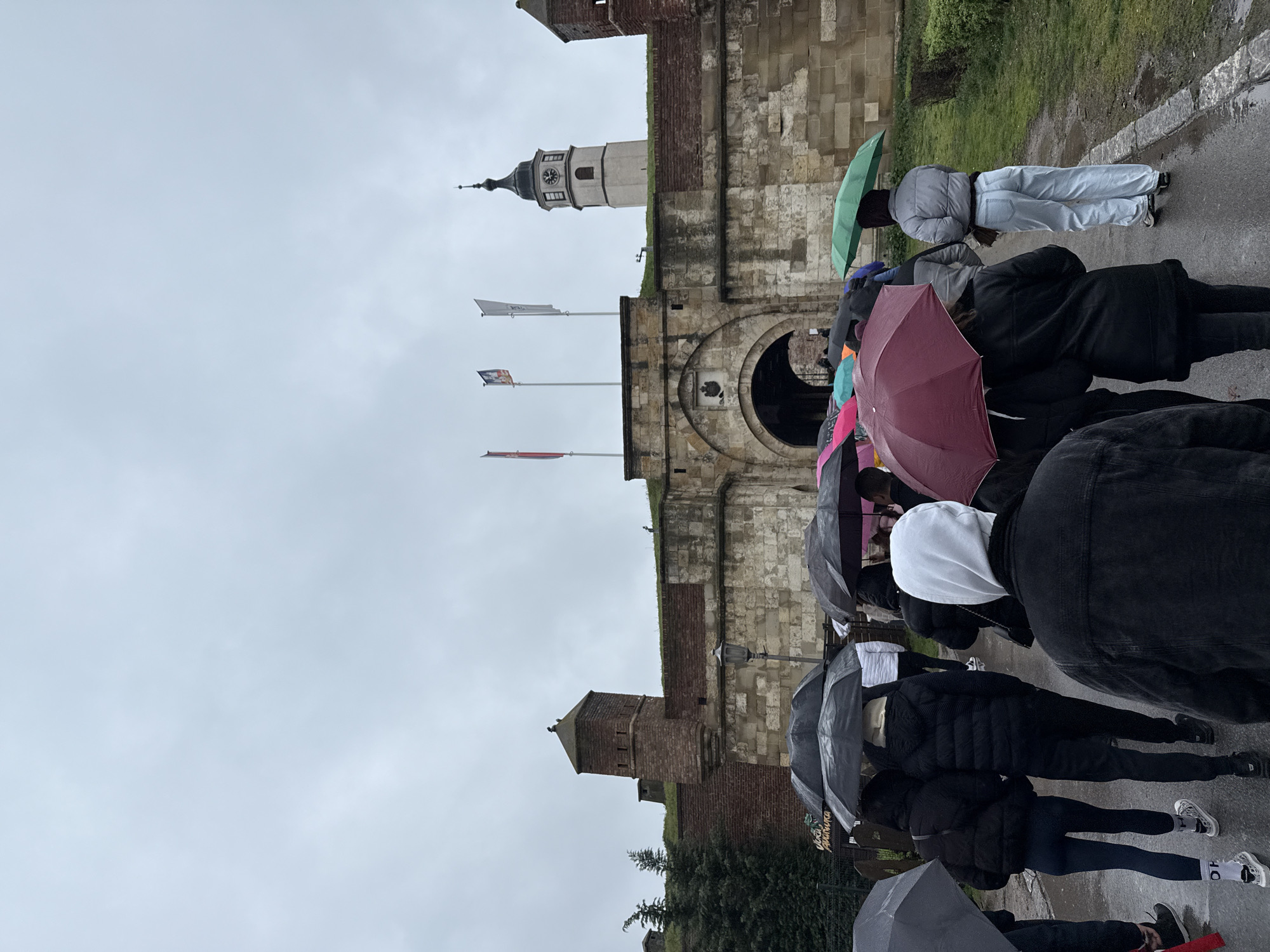 The image displays a group of multiple pedestrians walking on a wet paved pathway towards the Stambol Gate of the Belgrade Fortress in Belgrade, Serbia, under an overcast, grey sky. Most individuals are seen from behind, holding open umbrellas of various colors, including black, dark red, green, light blue, and pink, indicating recent or ongoing rain.
In the background, the historic Belgrade Fortress is prominent, featuring robust stone and brick fortifications. The Stambol Gate presents a large arched entrance topped with a carved stone emblem. To the left of the gate, a brick guard tower with slit-like openings rises above the wall, and another portion of a similar tower is visible on the far right. Above the fortress walls, the distinct white Sahat Kula (Clock Tower) with a dark, pointed spire and visible clock faces stands to the right of the main gate. Three flags on poles are flying atop the fortress wall above the gate. Green grass and sparse vegetation line the upper parts of the walls and the edges of the pathway. A black metal fence and a sign with Cyrillic text are partially visible on the left side of the path.