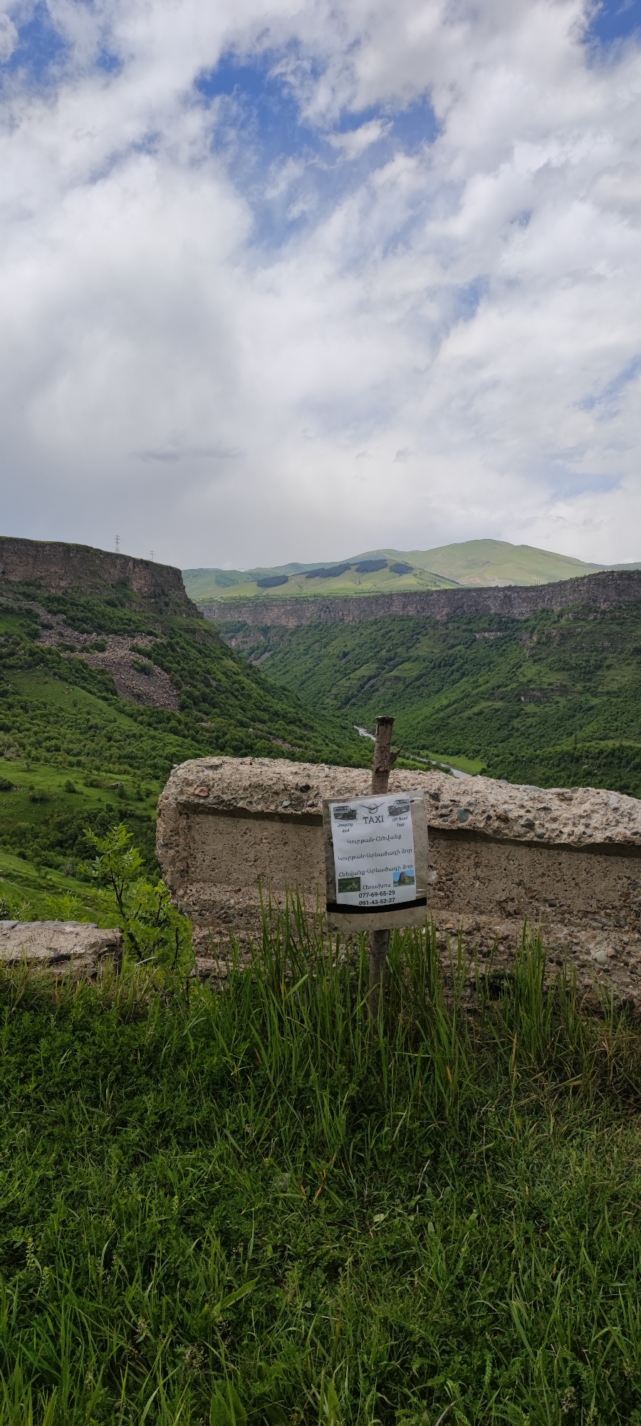 The image depicts a scenic view of a valley in Kurtan, Armenia. The foreground features a concrete structure partially obscured by tall grass. Affixed to this structure is a sign advertising taxi services, specifically mentioning routes to Hnevank Monastery and Arevatsag Gorge, both popular destinations in the area. The landscape consists of steep, green hills leading to a canyon. The sky above is filled with scattered clouds.