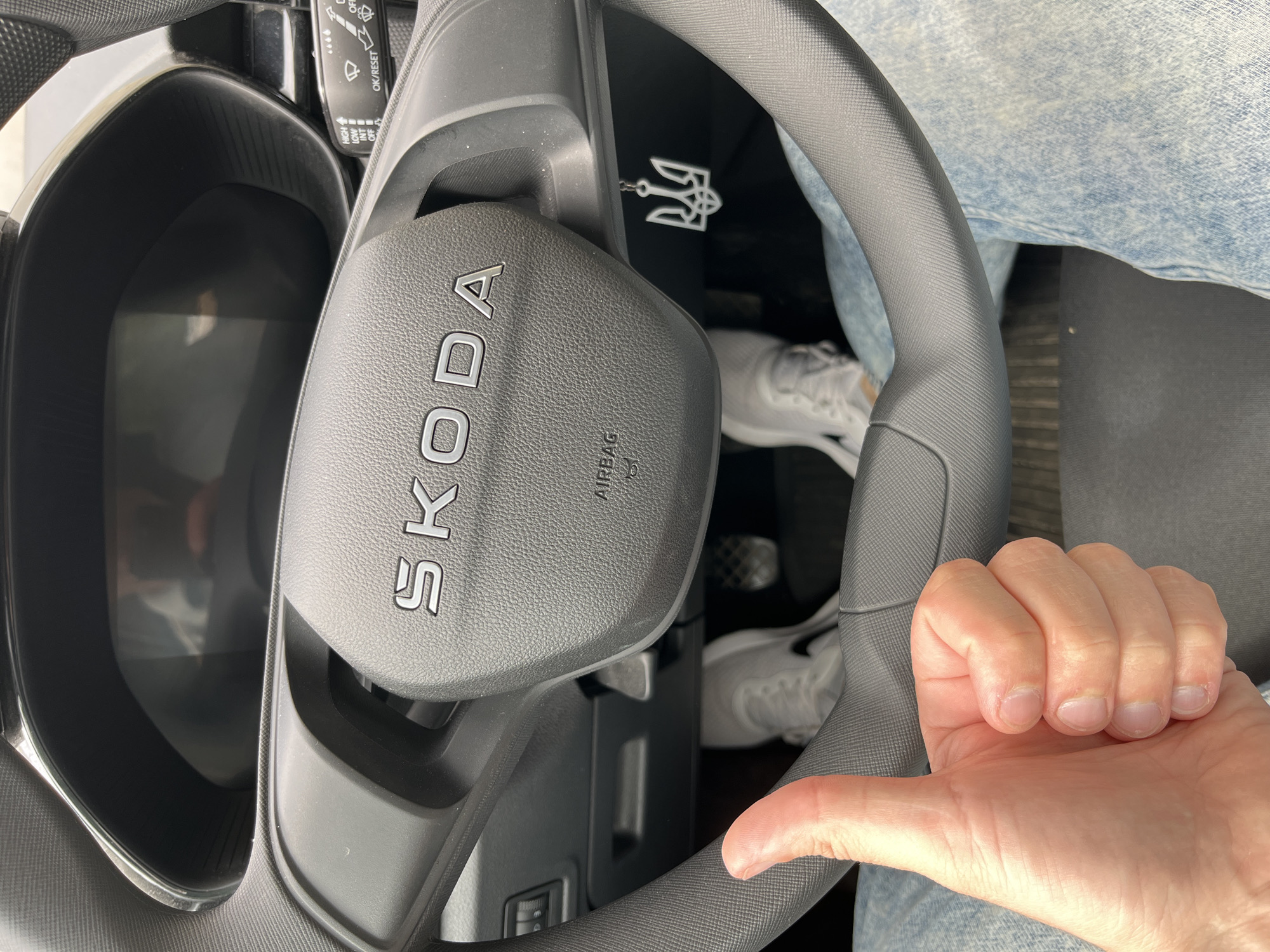 A first-person perspective shot shows the interior of a car, focusing on the steering wheel and dashboard. The steering wheel has the "SKODA" logo embossed on it and the "AIRBAG" indicator. A white keychain shaped like a trident, a symbol of Ukraine, hangs from the steering column. The driver's left hand is giving a thumbs-up, and the person is wearing light blue jeans and white sneakers. The car's pedals are visible in the foreground, along with a glimpse of the driver's side floor mat. The background shows the car's digital dashboard, which is dark, and parts of the car's interior. The image was taken inside a vehicle, likely parked, with no specific location in Kharkiv, Ukraine, identifiable.
