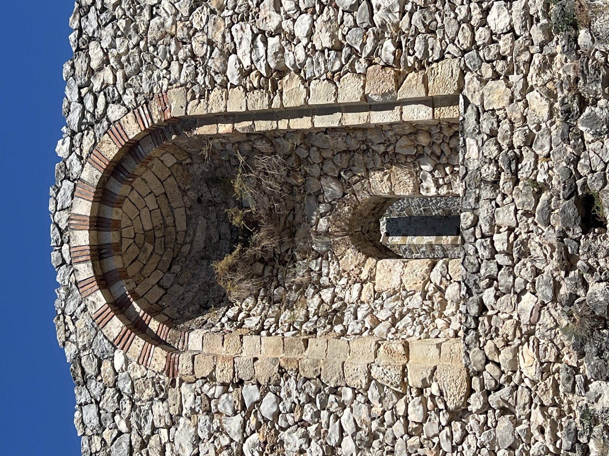 The image shows a close-up, eye-level view of an ancient stone wall, possibly part of a historical fortification, with a clear blue sky in the background. The wall is constructed from irregular, roughly cut stones of varying sizes and colors, predominantly in shades of grey, beige, and brown, with some white and pinkish hues. The stones are tightly packed, creating a rugged texture.
Prominently featured is a large arched recess built into the wall. The arch itself is constructed with alternating voussoirs of lighter-colored stone and reddish-brown brick, giving it a striped appearance. Within this large recess, there is a smaller, rectangular opening, also framed with stones, containing a dark interior suggesting it is a window or a narrow passage. Dried vegetation, resembling straw or dry grass, is lodged within the upper part of the large recess. The stonework around the smaller opening and the sides of the main recess uses more uniform, squared-off blocks of light beige stone.
The overall impression is one of age and a well-preserved historical structure. The setting appears to be outdoors, under natural sunlight, casting subtle shadows that highlight the texture of the stonework. This wall is located at the Despotovac Monastery, specifically the Church of St. Stephen, near Despotovac Municipality in Serbia.