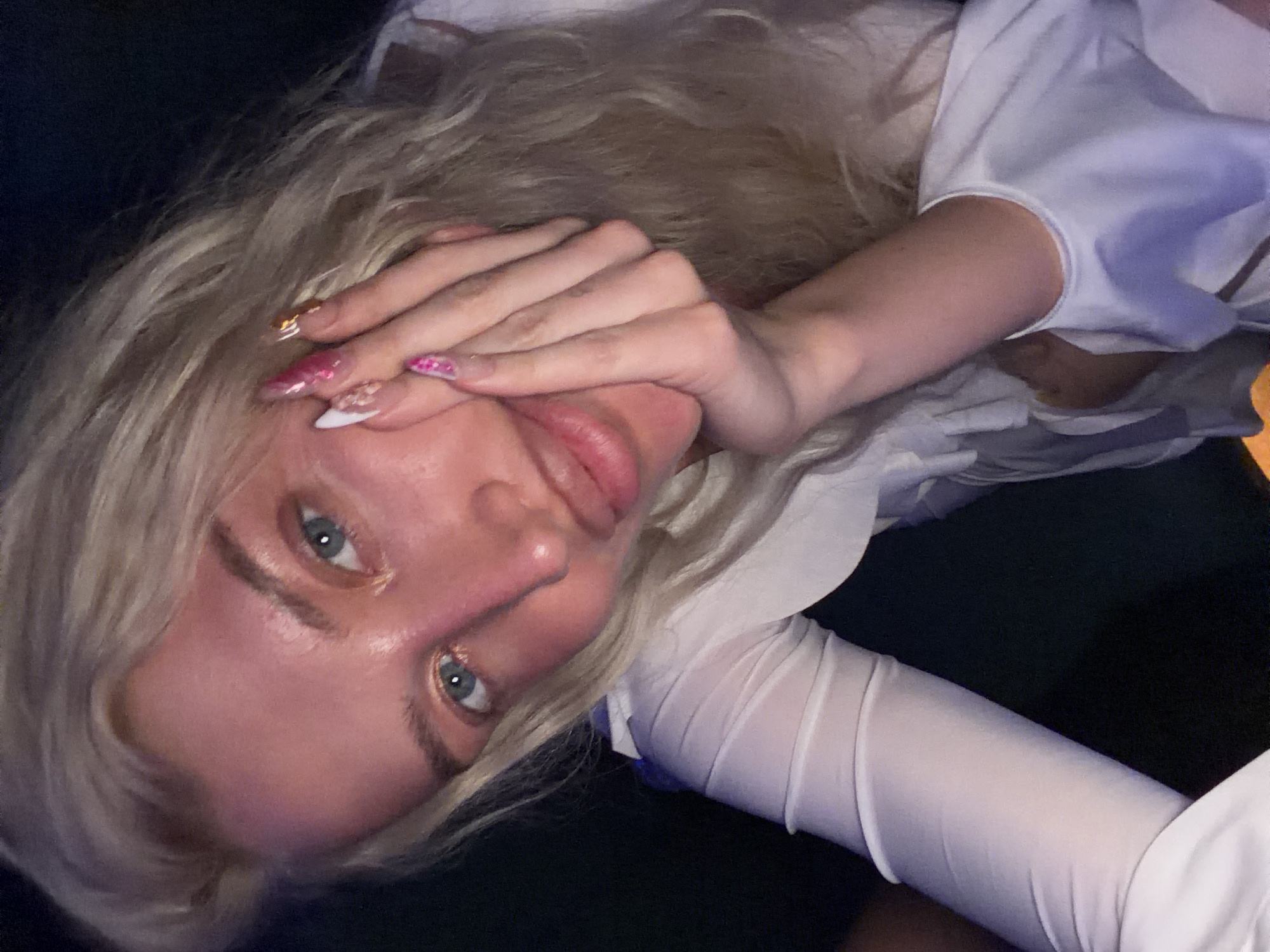 The image captures a close-up, rotated view of a person with long, wavy blonde hair and striking blue-green eyes, looking directly at the camera. The person's fair skin shows some redness, particularly around the nose and cheeks. One hand is raised, partially covering the lower part of the face and mouth, with fingers delicately posed.
A notable detail is the elaborate manicure on the visible hand. The nails are long and pointed, adorned with intricate designs featuring pink, white, and gold glitter, as well as small embellishments. The person is wearing a white top, possibly a shirt, with visible sheer fabric on the sleeves.
The setting appears to be dimly lit, with a dark background suggesting an indoor environment or nighttime. The lighting highlights the person's face and hair, creating some subtle reflections. There is no visible text in the image. The pose and dim lighting lend a somewhat intimate and contemplative mood to the photograph. No specific location cues are visible within the image itself.