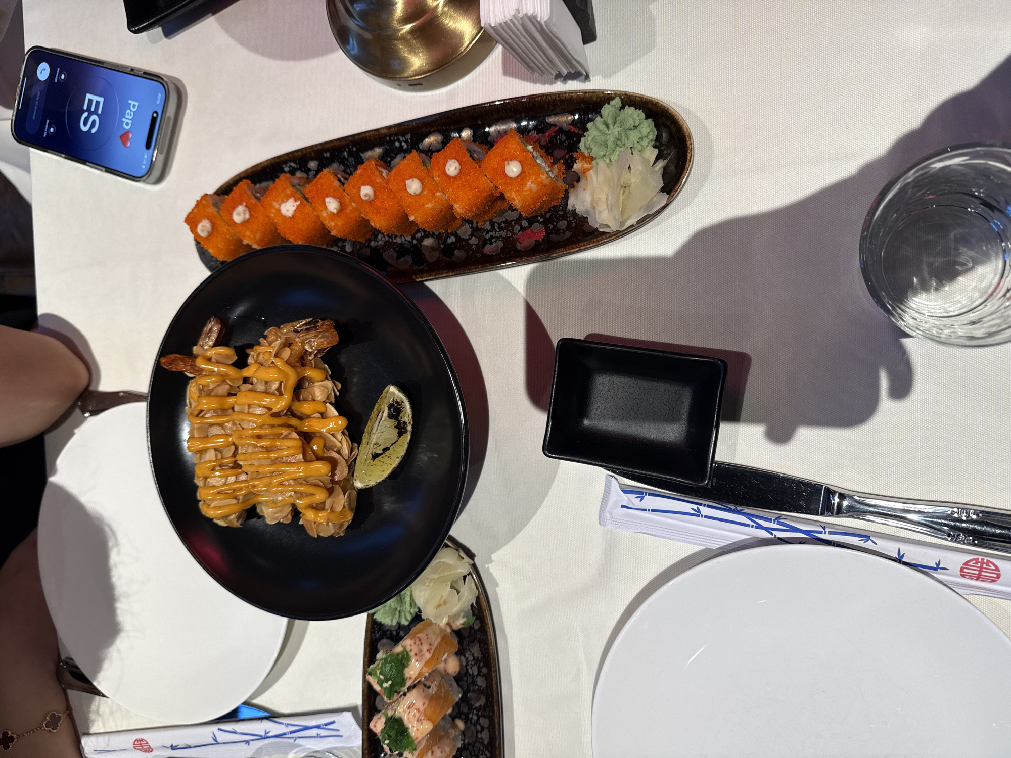 The image captures a dining table in a restaurant, possibly located in Yerevan, Armenia. The table is covered with a white tablecloth.
On the table are various dishes including sushi rolls arranged on a dark-colored plate, a plate with shrimp covered in a sauce, and a small plate with salmon sushi. There is also a small black dipping dish. Chopsticks in a wrapper and cutlery are placed near a white plate. A glass of water is also visible.
A smartphone, displaying an incoming call labeled "Pap" with a heart icon next to it, rests on the table beside the sushi. The lighting suggests an indoor setting. A person's arm, adorned with a clover bracelet, is visible in the lower left corner of the image.