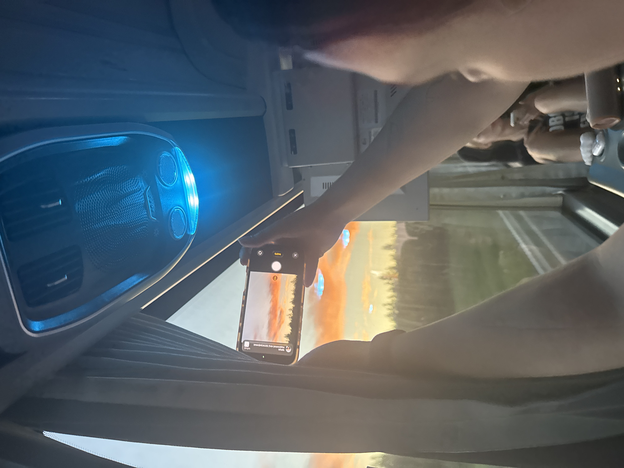 A person in Федькино, Russia, is taking a photo with their smartphone of the landscape outside the window of a vehicle, possibly a bus or van. Other passengers are visible in the background. The interior of the vehicle includes a blue-lit ceiling console.