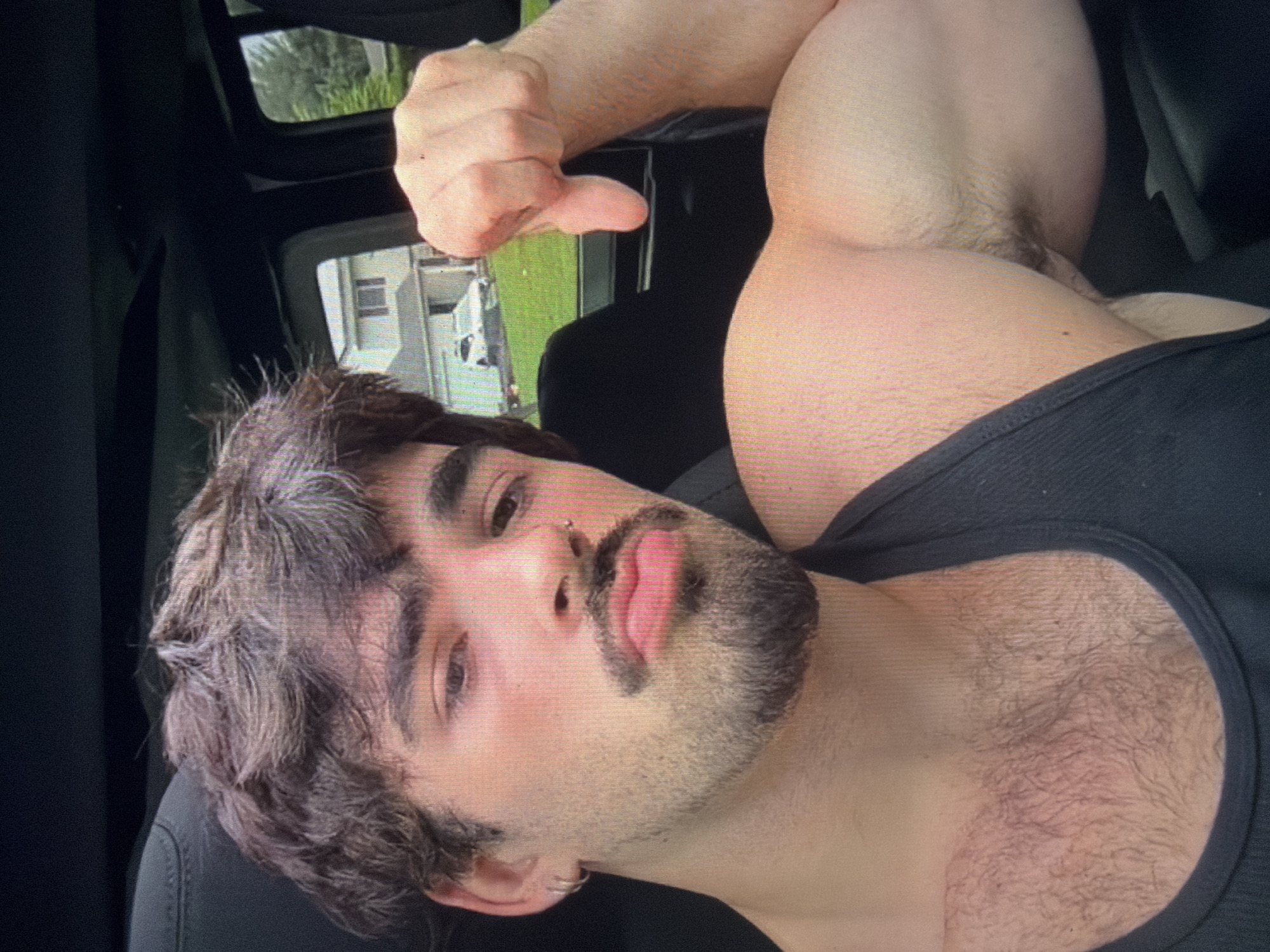 A male individual with dark, wavy hair, a dark mustache, and a beard is depicted in a selfie taken from inside a vehicle. He wears a dark tank top, a silver stud nose piercing on his right nostril, and a silver hoop earring on his left ear. His right arm is flexed, showcasing a well-developed bicep, with his hand curled into a fist, thumb pointing downwards. Visible chest hair is present beneath his collar. Through the vehicle's window, a residential area is observed, featuring a two-story house with dark windows, green lawn, and foliage, along with a white SUV parked on a paved surface. This scene is located in the city of Shumen, Bulgaria.