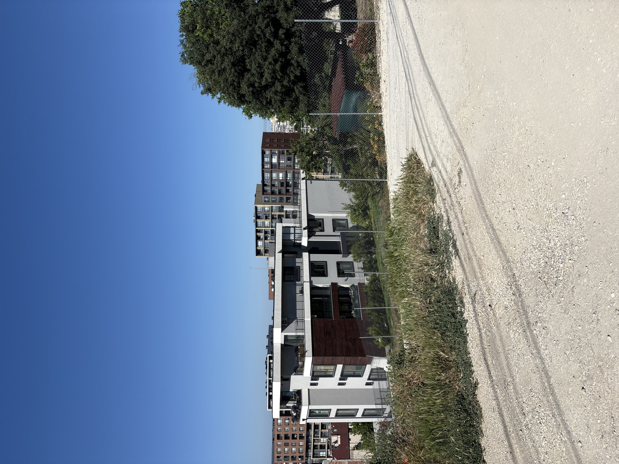 A bright, clear, sunny day in Varna, Bulgaria, with a wide shot of a modern apartment complex in the foreground, set against a backdrop of other buildings and a lush green tree. The foreground features a dusty, gravelly path with tire tracks, leading up a gentle incline. Tall grasses and other vegetation grow along the edge of the path, obscuring a chain-link fence.
The apartment building is a multi-story structure with a grey and white facade, accented by a section of dark wood paneling. It has multiple balconies with railings and large windows. Other buildings, varying in height and architectural style, are visible in the middle ground and background, suggesting an urban or suburban environment. The sky is a vibrant, cloudless blue. A large, mature tree with dense foliage is situated to the right of the apartment complex. The scene suggests a residential area under development or undergoing renovation, given the presence of the undeveloped path and fencing. There are no people visible in the image.